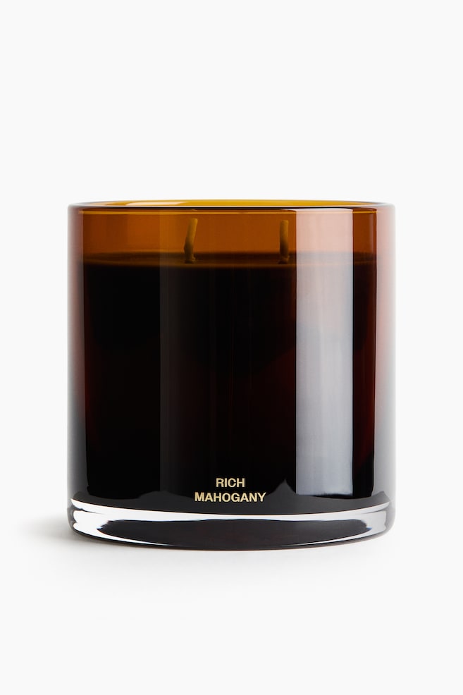Een geurkaars in een cilindrische amberkleurige glazen houder staat naar voren gericht, met donker was en twee zichtbare lonten. Aan de voorzijde onderaan van de glazen container staan de woorden "RICH MAHOGANY" in kleine gouden hoofdletters boven een dikke, transparante voet. - 1