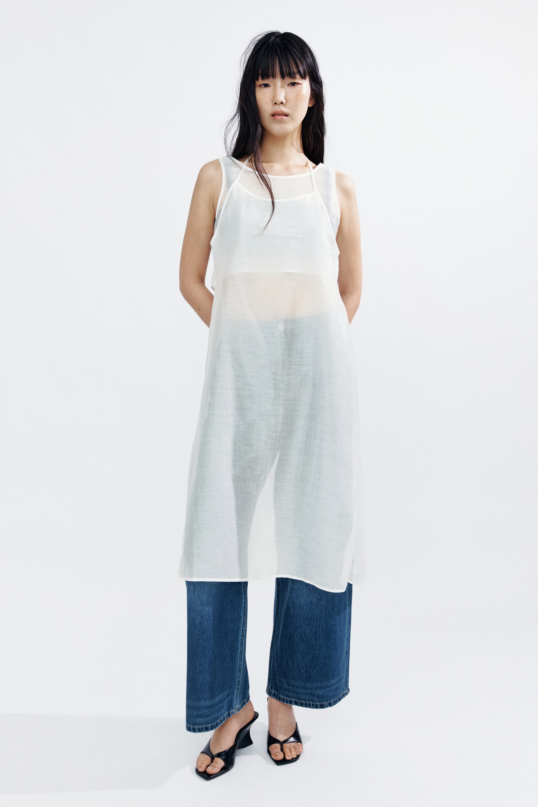拡大画像を表示: モデルが着用しているのは、ふくらはぎ丈のシアーホワイトドレスです。ラウンドネックラインと細いショルダーストラップが特徴で、ダークなクロップドトップの上に重ねられています。この裏地のないドレスは、ブルーのワイドレッグジーンズとブラックのオープントゥサンダルとスタイリングされています。