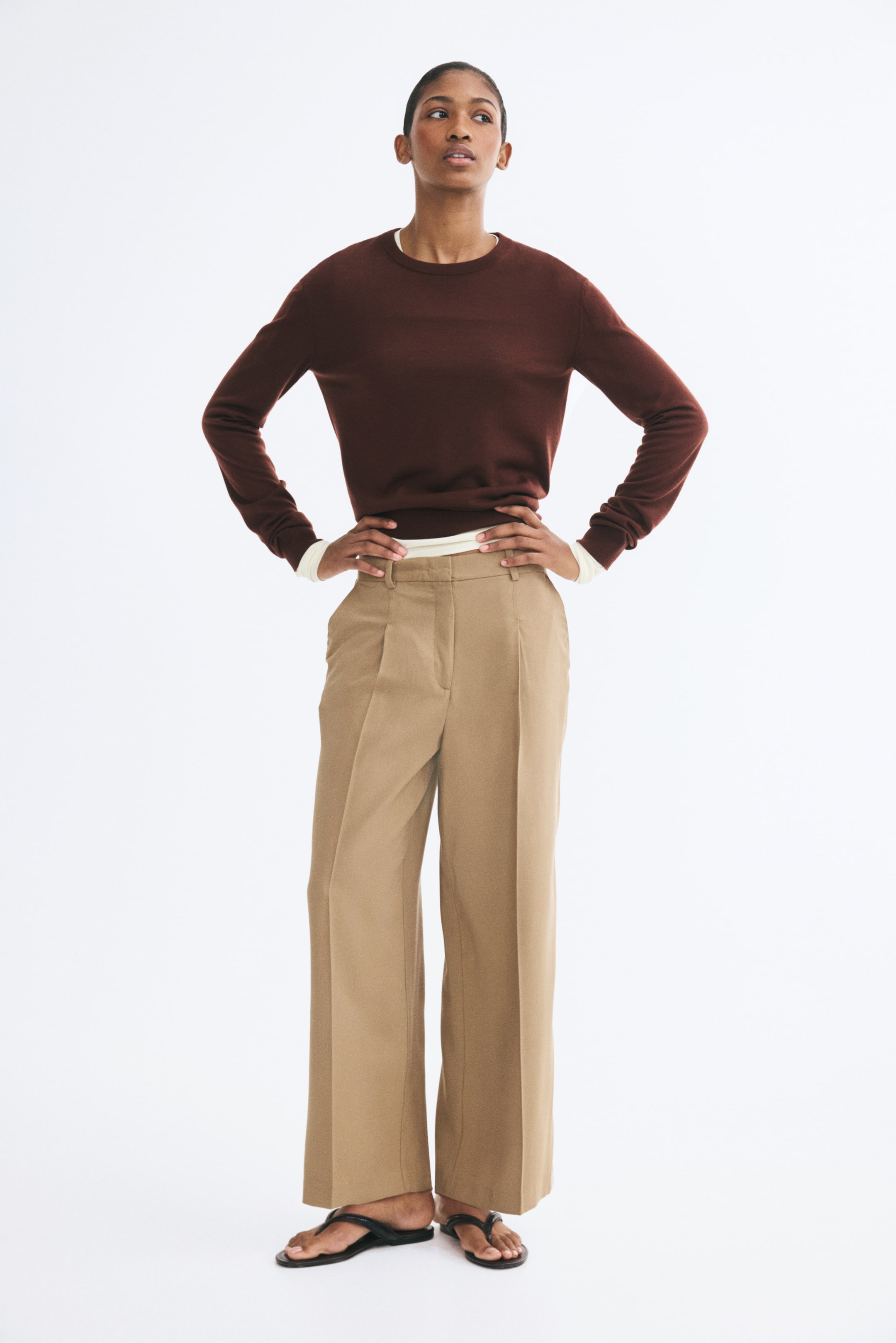 Ampliar la imagen: Pantalones beige hasta el tobillo con cintura alta, piernas rectas con líneas marcadas al frente y bolsas laterales diagonales, llevados por la modelo. La modelo está de pie con las manos en las caderas, combinando los pantalones con un suéter café de cuello redondo y sandalias planas negras.