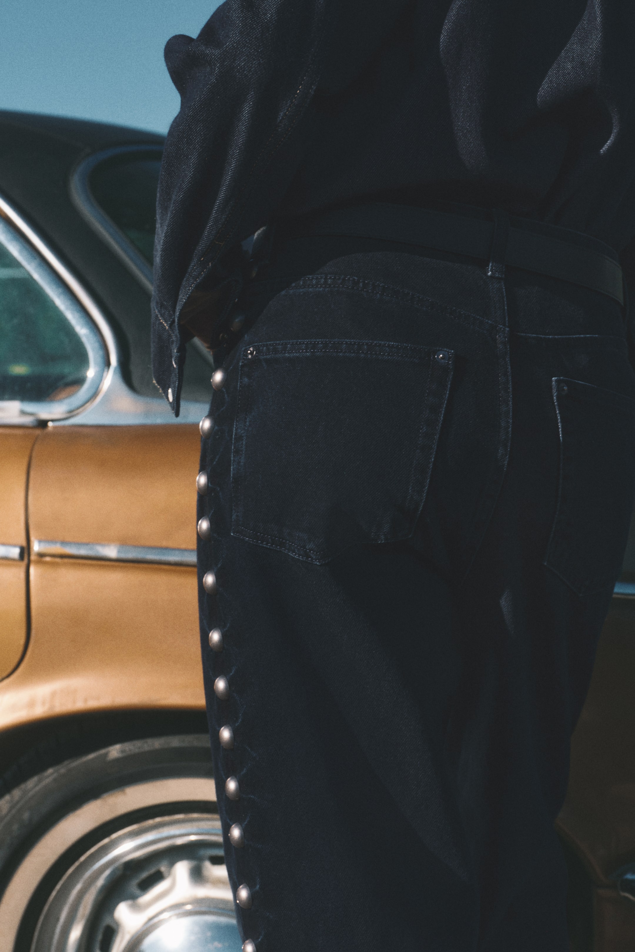 Voir une image plus grande: Un jean en denim de lavage foncé est présenté du côté arrière gauche, avec des poches plaquées à l'arrière et une ligne verticale de clous métalliques ronds le long de la couture extérieure de la jambe gauche. Une ceinture foncée est portée avec le jean, qui est assorti à une veste en denim de lavage foncé.