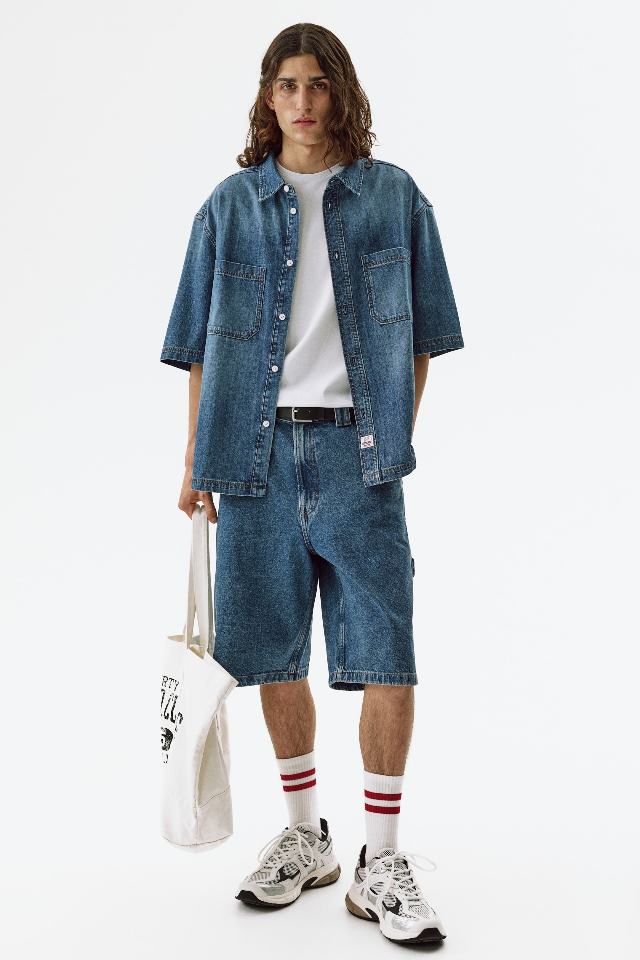 View larger image: The product showcases a complete outfit facing forward, highlighting a faded blue denim short-sleeve button-up shirt with two chest patch pockets, layered over a white crew-neck t-shirt, and matching faded blue denim knee-length shorts paired with a black belt. The ensemble is finished with white mid-calf socks featuring red stripes, chunky athletic sneakers, and a cream canvas tote bag with black graphic text.