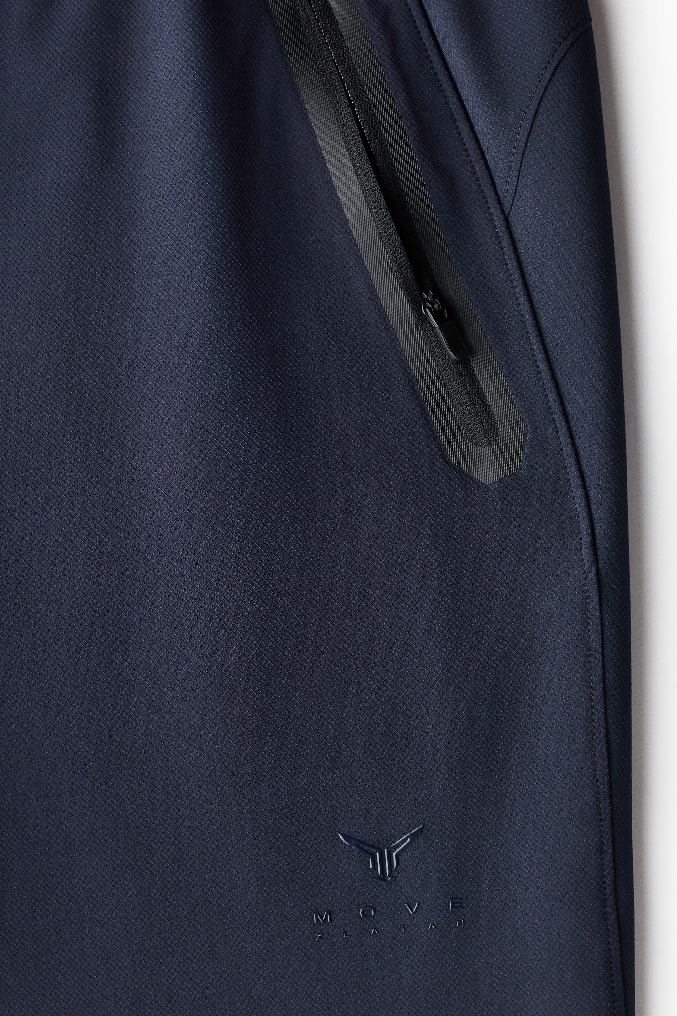 Ampliar la imagen: Una prenda deportiva azul marino, vista desde el FRENTE, presenta un tejido con textura de cuadrícula sutil. Un bolsillo con cierre negro en ángulo, con un tirador negro, está posicionado en la parte superior derecha, y un logo en relieve tono sobre tono 'MOVE ZLATAN' aparece en la parte inferior izquierda.