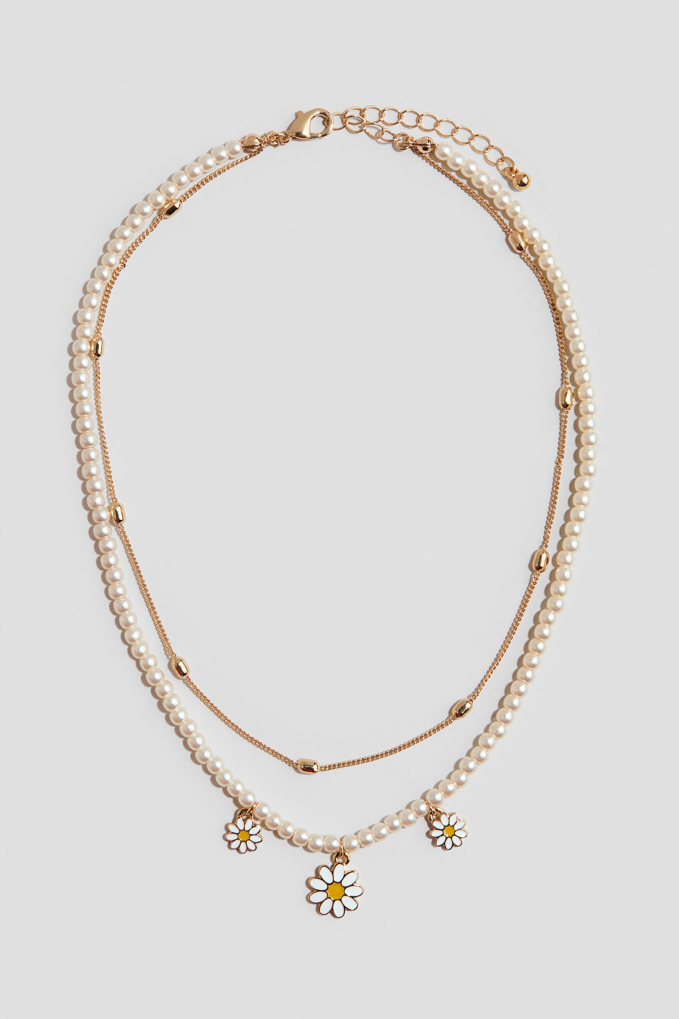 Agrandir l'image: Un collier en couches, présenté de face, se compose de deux rangées : l'une de petites perles couleur crème et l'autre d'une fine chaîne dorée avec des perles dorées intermittentes. Trois breloques en forme de marguerite avec des pétales blancs et des centres jaunes pendent de la rangée de perles, tandis qu'un fermoir mousqueton doré et une chaîne d'extension sont visibles en haut.