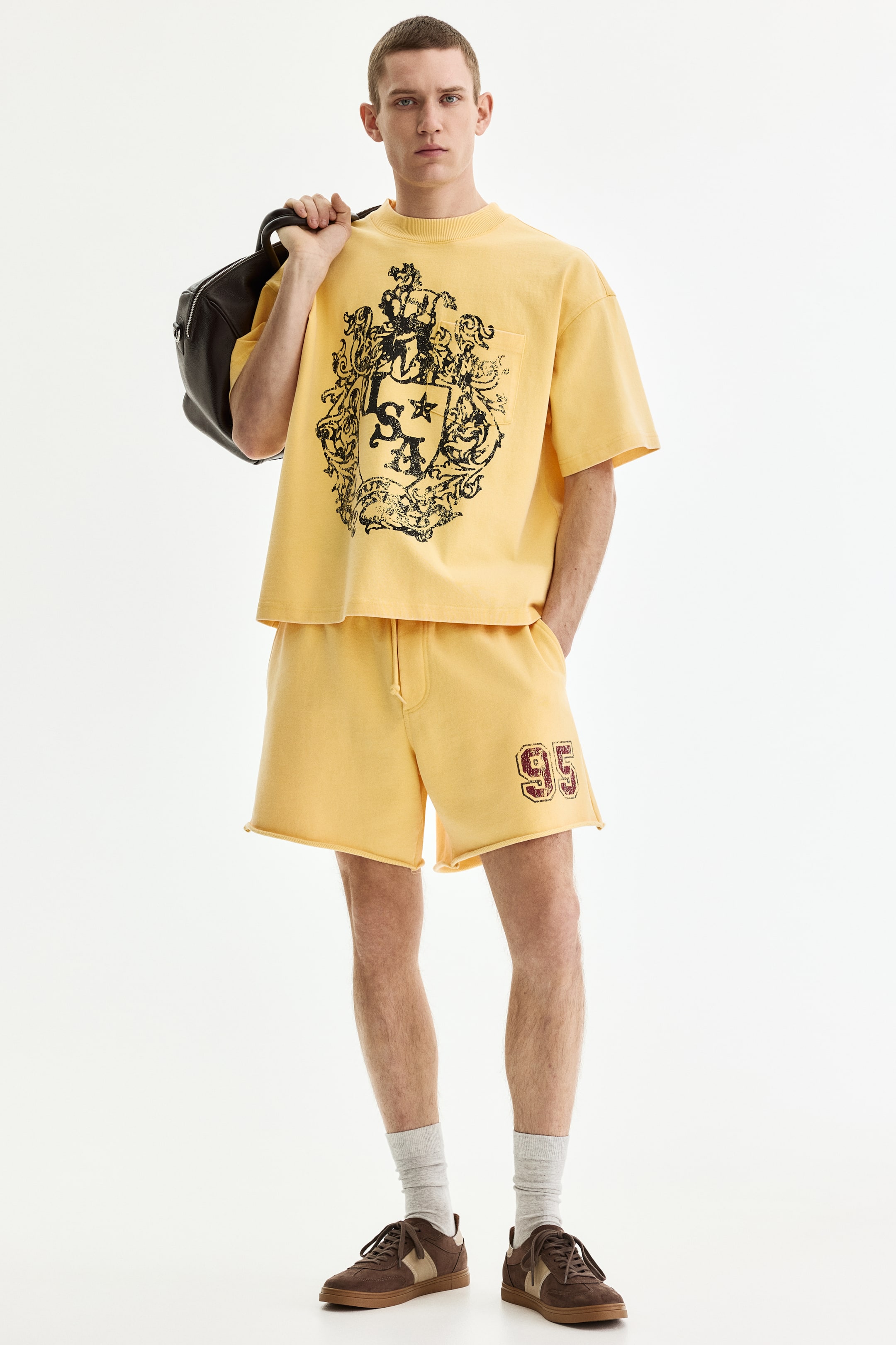 View larger image: A yellow T-shirt and matching shorts set is shown front-facing. The oversized T-shirt features a ribbed crewneck and a large black crest graphic on the front, while the shorts feature a drawstring waist, raw-cut hems, and a burgundy "95" graphic on the left thigh.