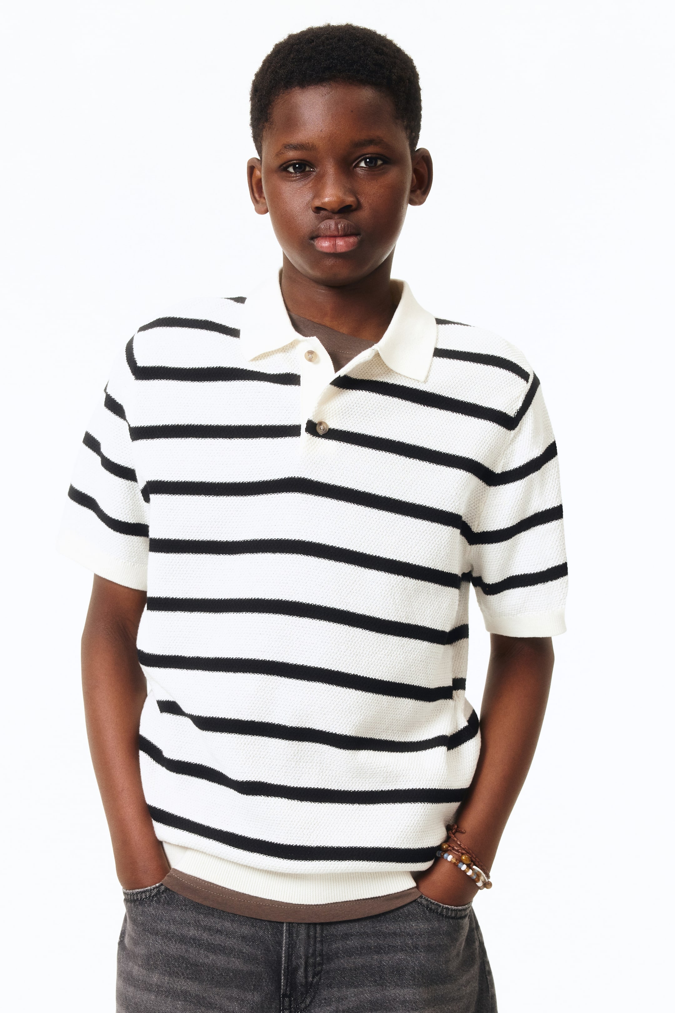 Ampliar la imagen: La camisa polo blanca con rayas horizontales negras muestra una tela de punto texturizada. Presenta un cuello clásico blanco, una tapeta de dos botones con botones marrón claro, mangas cortas y un dobladillo acanalado.