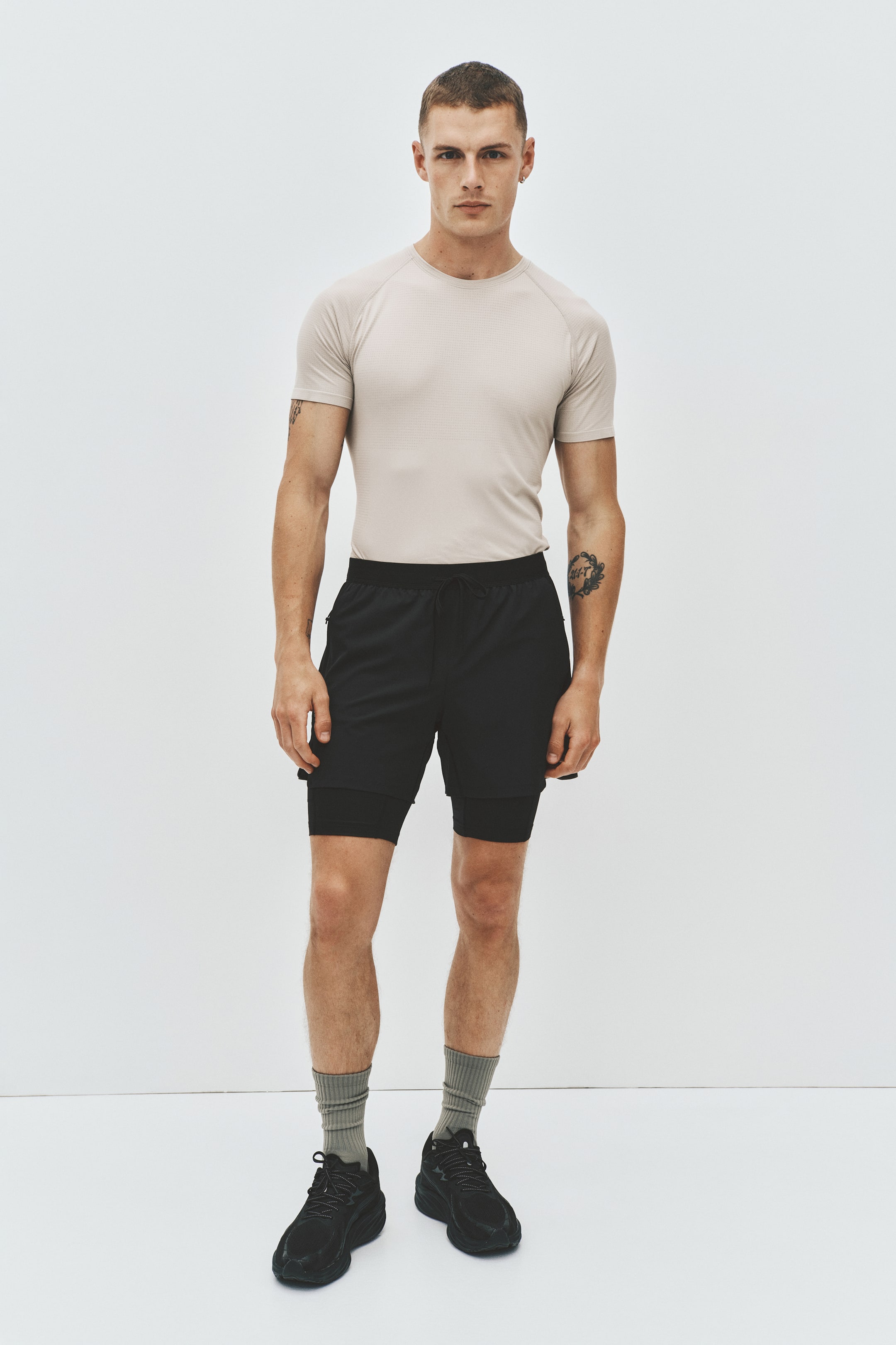View larger image: A light beige crew-neck t-shirt with raglan sleeves and subtle textured fabric is being worn, paired with black layered shorts featuring an elastic drawstring waistband, side zippered pockets, and a longer inner compression layer. Black athletic shoes with chunky soles are completing the outfit, worn with ribbed grey crew socks, all presented from a front-facing angle.