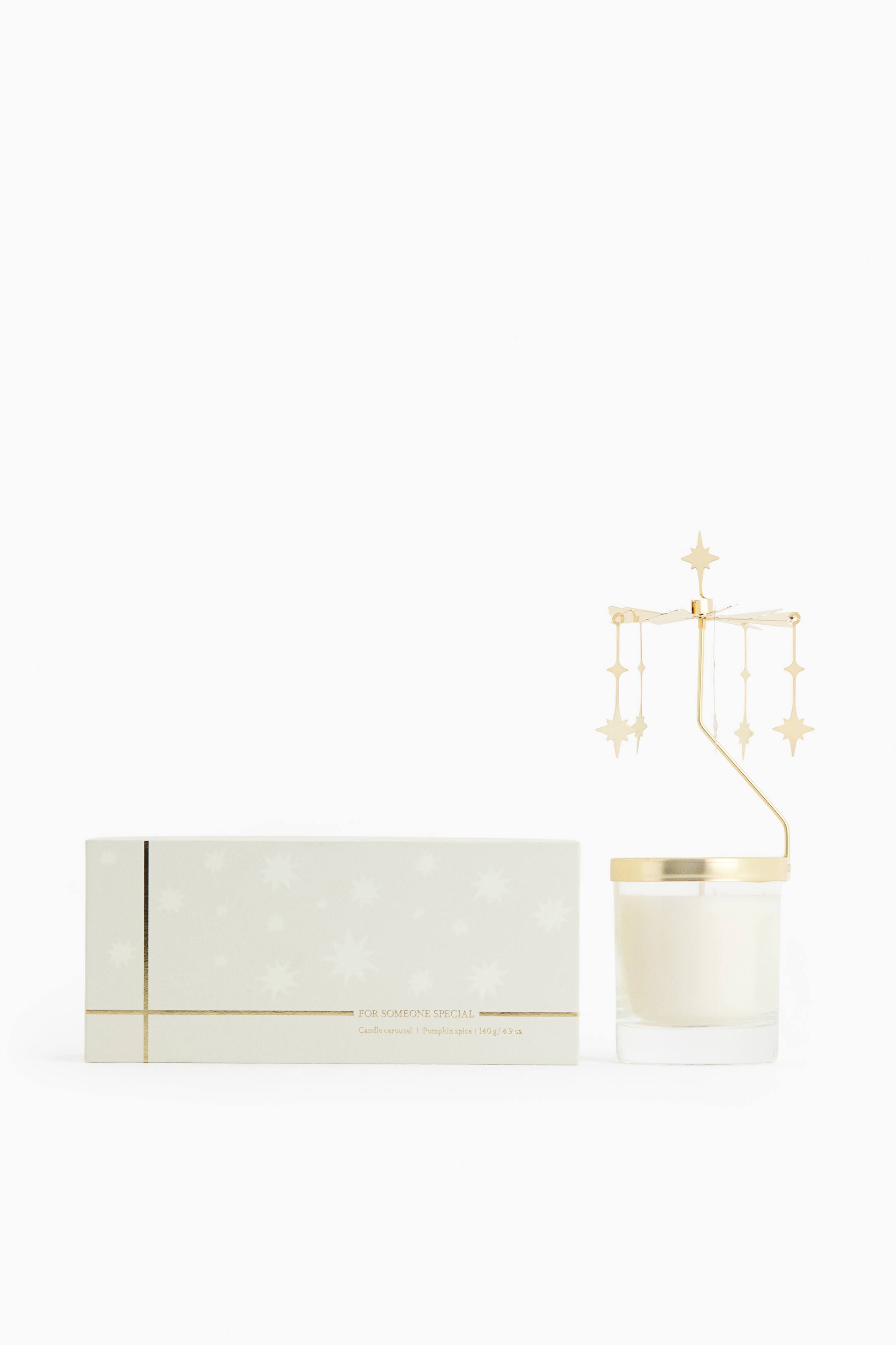 Ver imagen más grande: Un set de carrusel para velas, mostrado de frente, presenta una vela blanca en un tarro de vidrio transparente junto a su caja de embalaje rectangular de color claro. El accesorio de carrusel de color dorado reposa sobre el borde del tarro con colgantes de estrellas, mientras que la caja a juego luce líneas de estampado en lámina dorada y motivos de estrellas.
