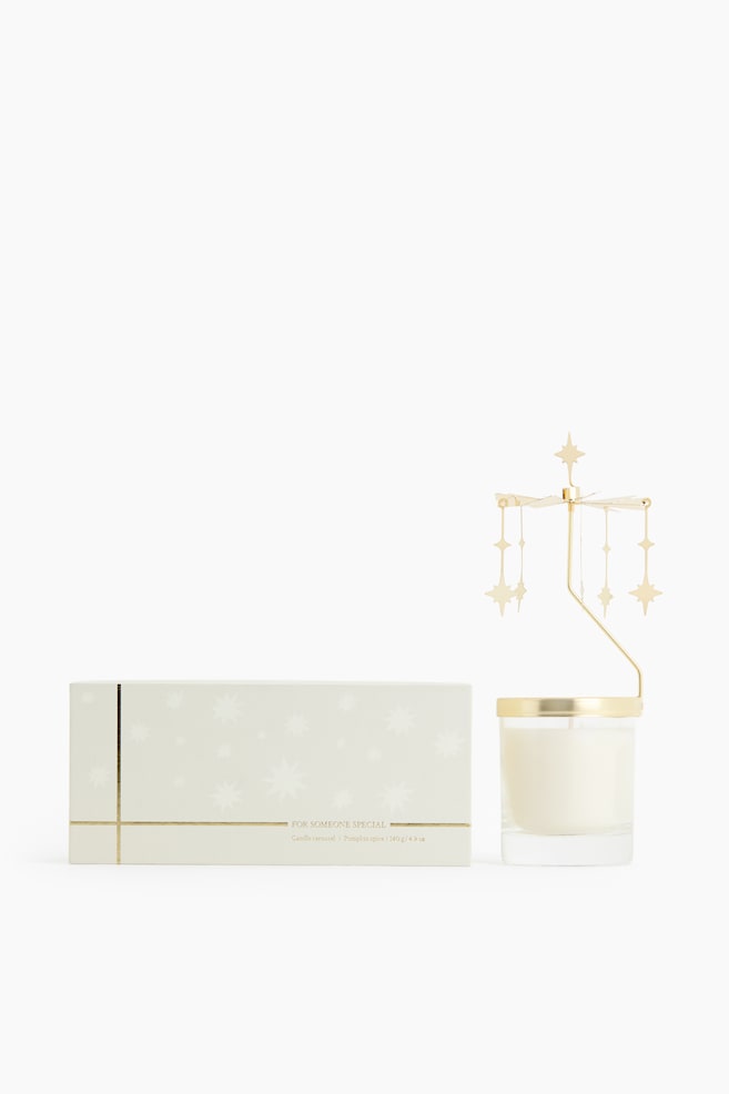 A candle carousel set, shown from the front, displays a white candle in a clear glass jar alongside its rectangular light-coloured packaging box. The gold-coloured carousel attachment rests on the rim of the jar with dangling star charms, while the matching box features gold foil lines and star patterns. - 1