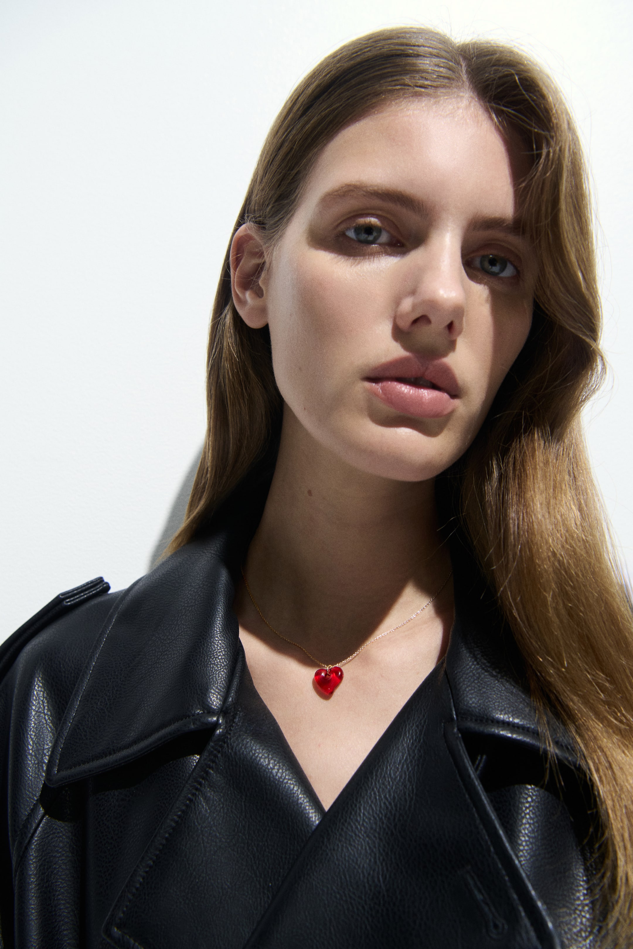 View larger image: A delicate gold-toned chain necklace displays a translucent red heart-shaped pendant. The pendant rests on the wearer’s décolletage, contrasting with a dark garment.