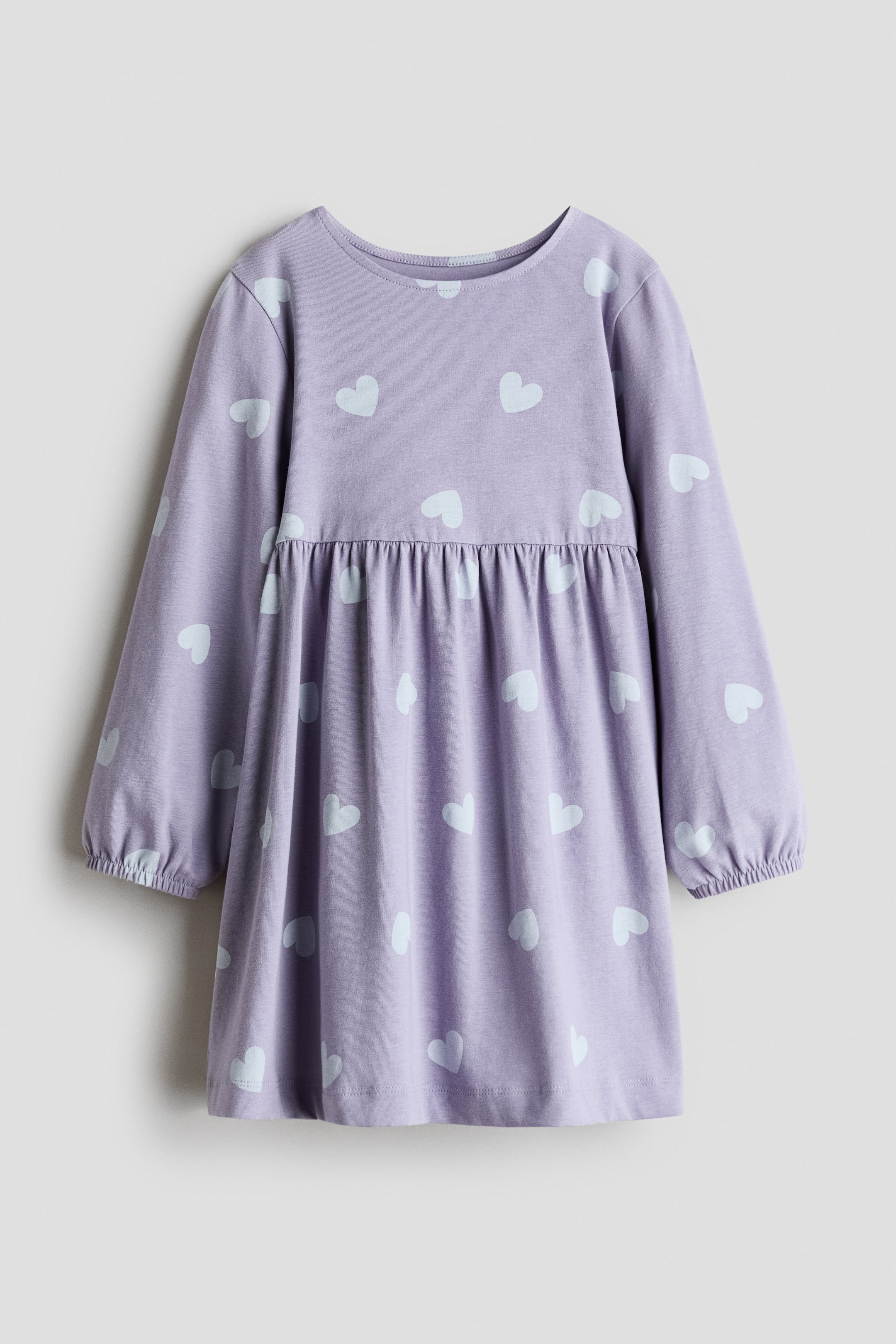 View larger image: A soft cotton jersey dress is shown from the front, showcasing a light purple hue adorned with a light blue heart pattern. It features a round neckline, long sleeves with elastic cuffs, and a gathered seam at the waist.