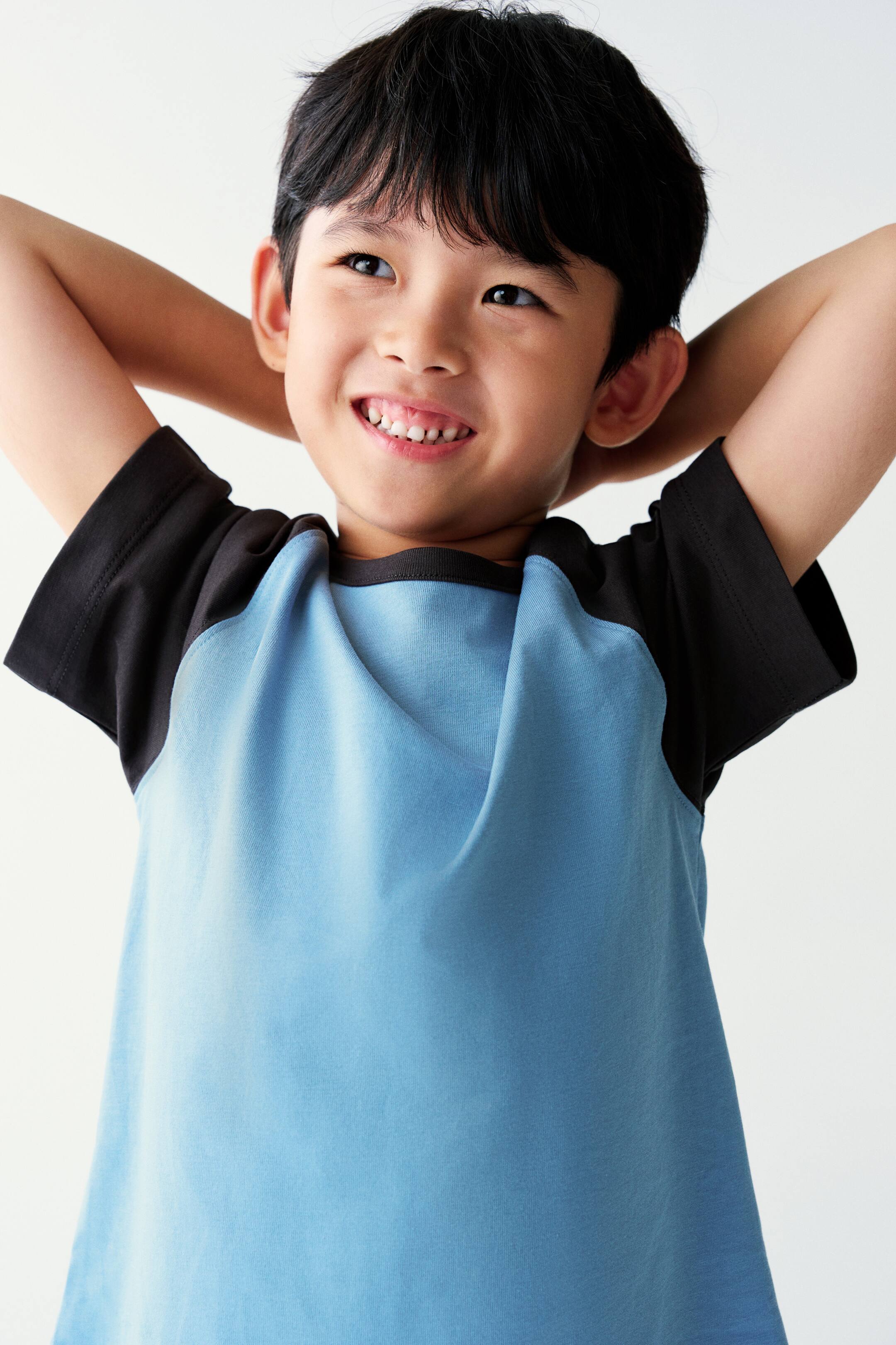 View larger image: A short-sleeved raglan T-shirt with a light blue body and contrasting black sleeves is worn by the model. The model is posing with hands placed behind the head, highlighting the crew neckline and fit of the shirt.