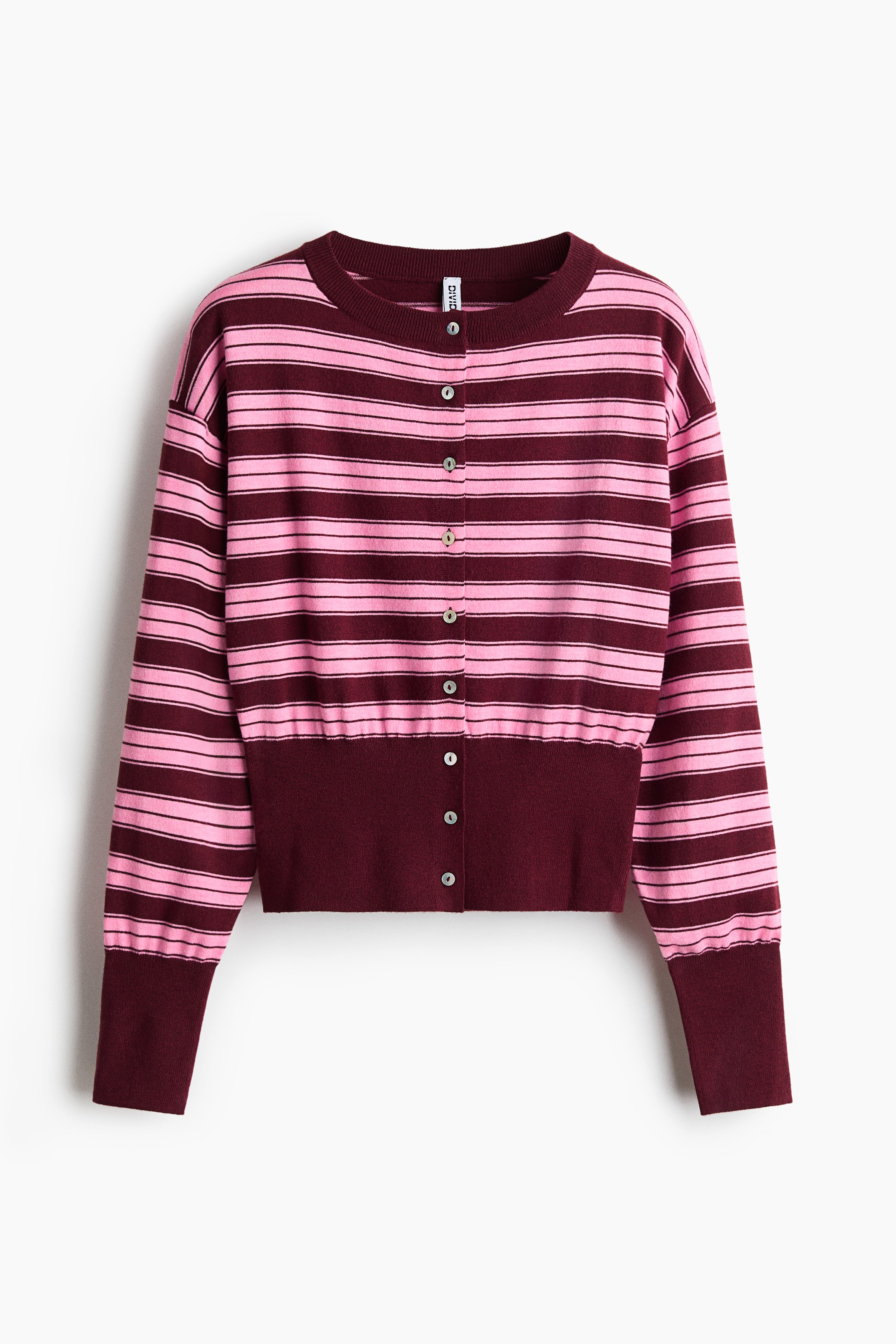 A short, fine-knit cardigan with pink and burgundy horizontal stripes is worn by the model, featuring a ribbed round neckline, button-front closure, dropped shoulders, and wide burgundy cuffs. The cardigan is styled with white trousers as the model sits with hands clasped over one knee.
