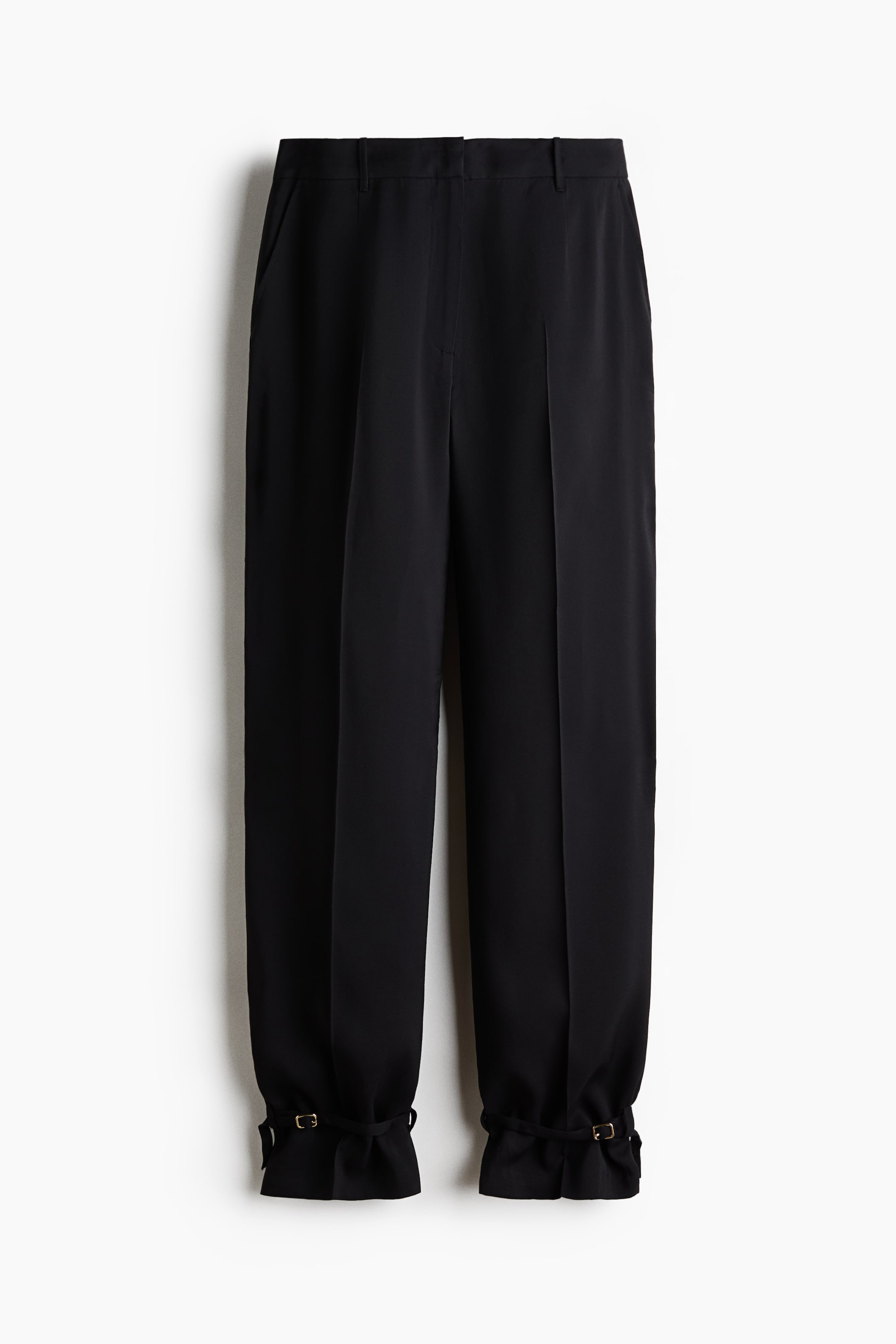View larger image: A pair of black trousers is shown from the front, featuring a relaxed fit with sharp center creases down the legs. The trousers taper at the hem with adjustable ankle straps and small gold-tone buckles, creating a gathered, cinched cuff effect.