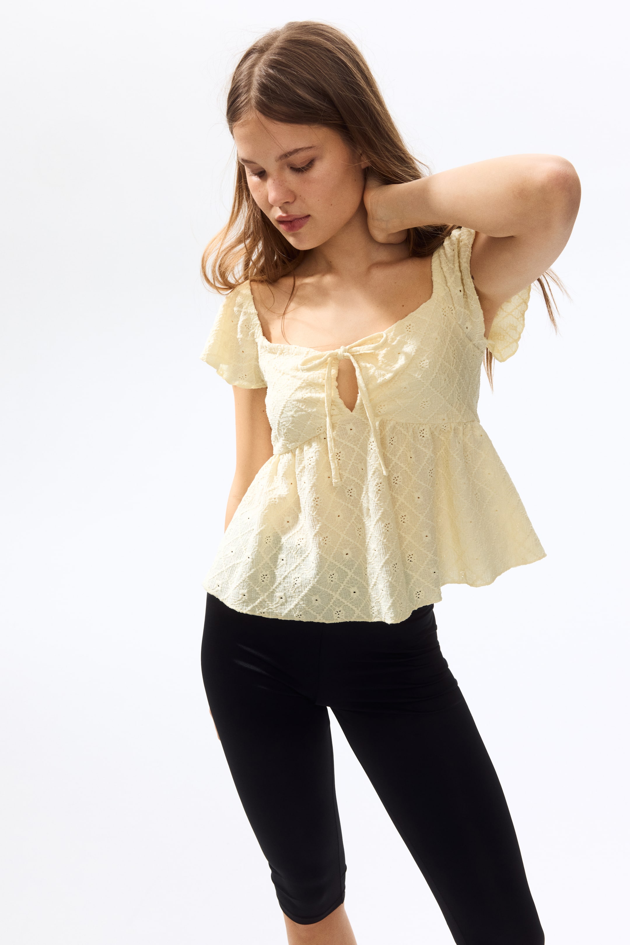 View larger image: A subtle yellow peplum top features a square neckline, short flutter sleeves, and intricate eyelet embroidery with a tie-front detail that forms a keyhole opening at the bust. This top is paired with black capri pants, highlighting its fit and length.