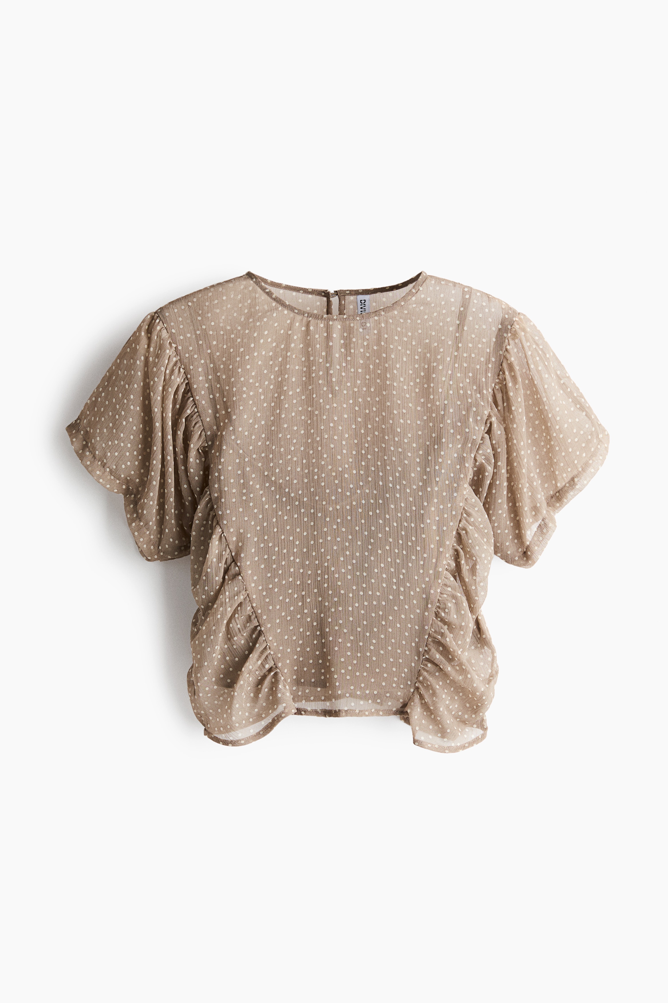 The model is wearing a sheer chiffon blouse in a taupe polka-dot print, featuring a round neckline, short flutter sleeves, and gathered side panels. The blouse is layered over a dark underlayer and paired with blue jeans.