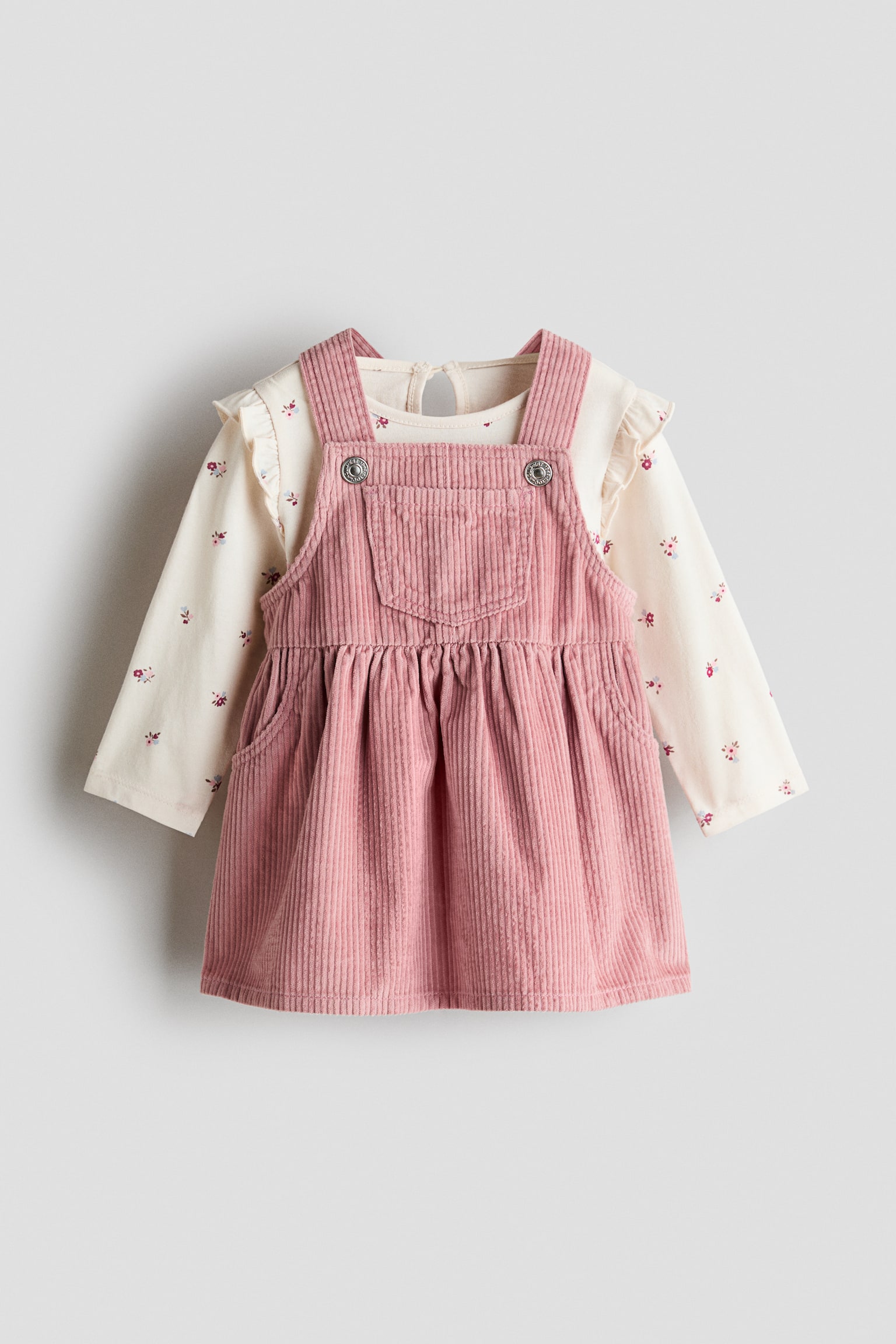 Ein rosa Cord-Pinaforekleid wird über einem cremefarbenen Langarmoberteil getragen und ist von vorne dargestellt. Das Pinaforekleid verfügt über Träger mit Knopfverschluss, eine mittige Brusttasche und einen gerafften Rock, während das Oberteil mit gerüschten Schulterdetails und einem zarten Blumenmuster präsentiert wird.