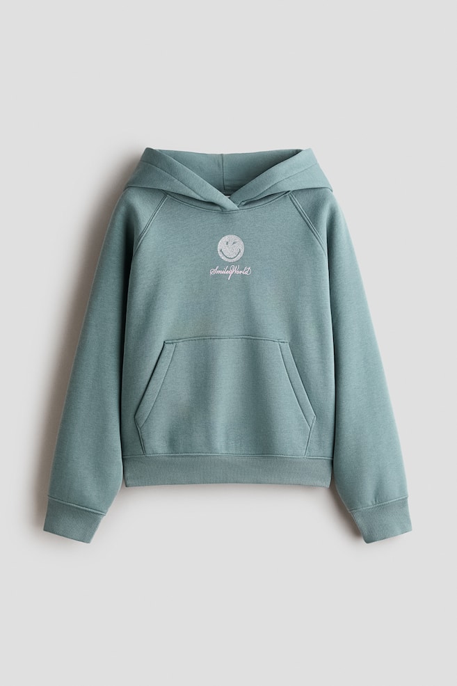 A front-facing view shows a dusty green oversized hoodie featuring a wrap-front hood, raglan sleeves, and a front kangaroo pocket. The chest is displaying a printed graphic of a winking smiley face with pink script text underneath, finished with wide ribbed trim on the cuffs and hem. - 1