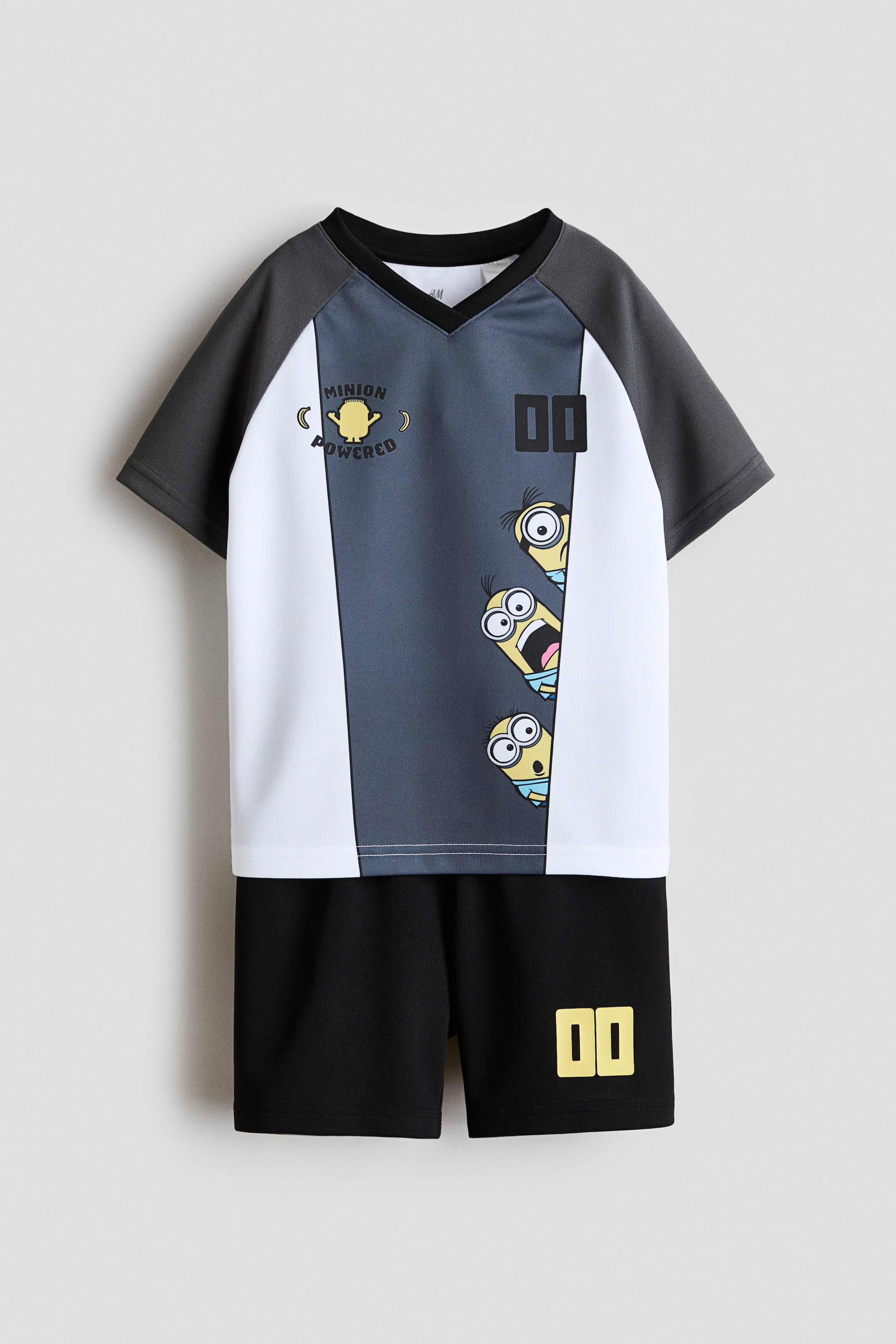Größeres Bild ansehen: Das Fußball-Set aus Oberteil und Shorts wird von vorne präsentiert. Das Oberteil zeigt weiße, dunkelgraue und mittelgraue Panels, einen schwarzen V-Ausschnitt mit Rippbündchen und dunkelgraue Raglanärmel. Es ist mit einem „Minion Powered“-Minion-Graphic, den Zahlen „00“ und drei hervorlugenden Minion-Figuren versehen. Die schwarzen Shorts zeigen passende „00“-Zahlen am unteren linken Bein.