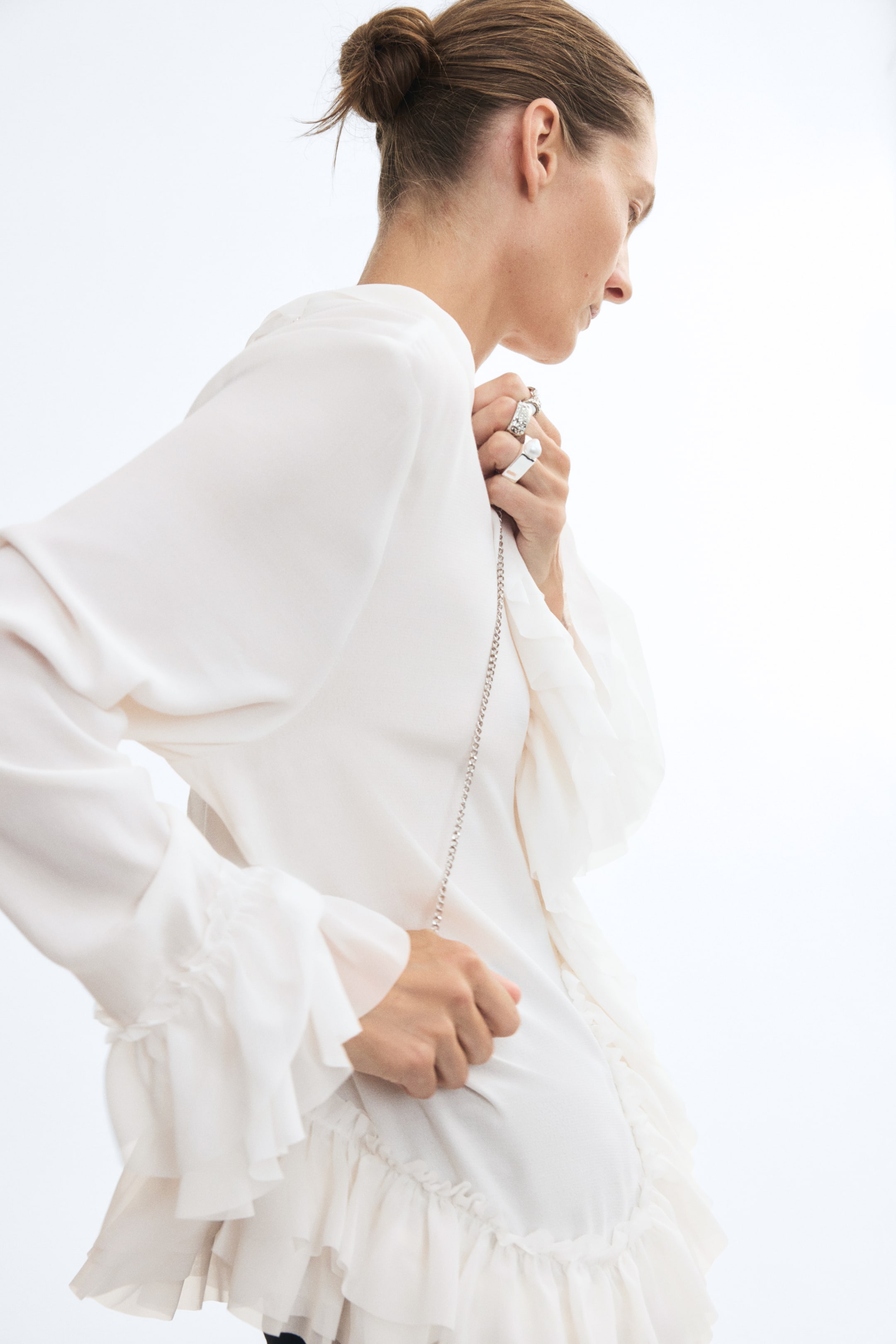 View larger image: The model is wearing a long off-white blouse in a lightweight, airy weave, featuring voluminous frill trims at the cuffs and hem. The model is holding a thin silver chain strap draped across the front of the blouse.