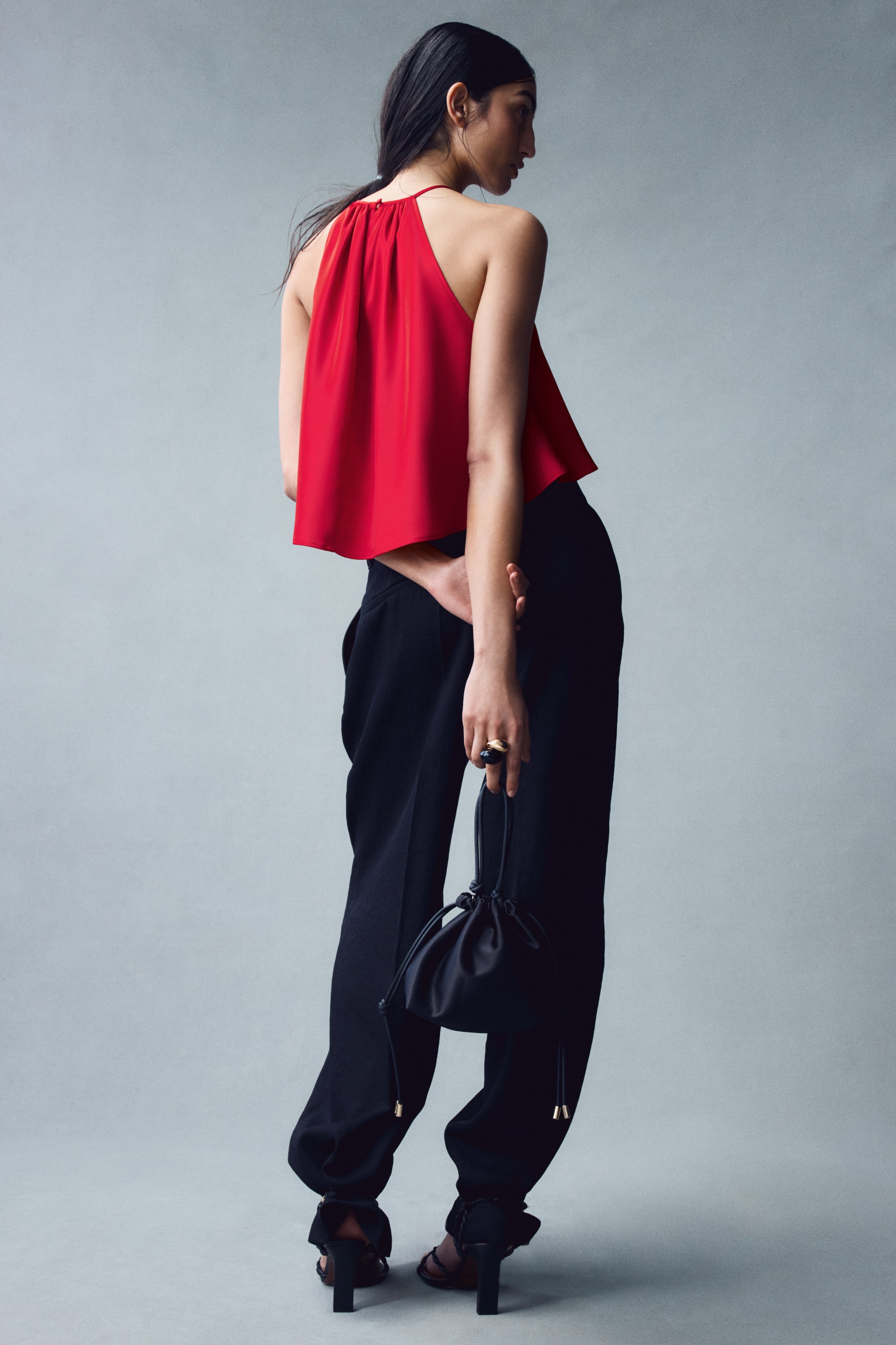 View larger image: The model is wearing a short, sleeveless red top featuring a high, gathered halter neckline and a loose, draped fit. The top is styled with black trousers, and the model is carrying a small black drawstring handbag behind their back.
