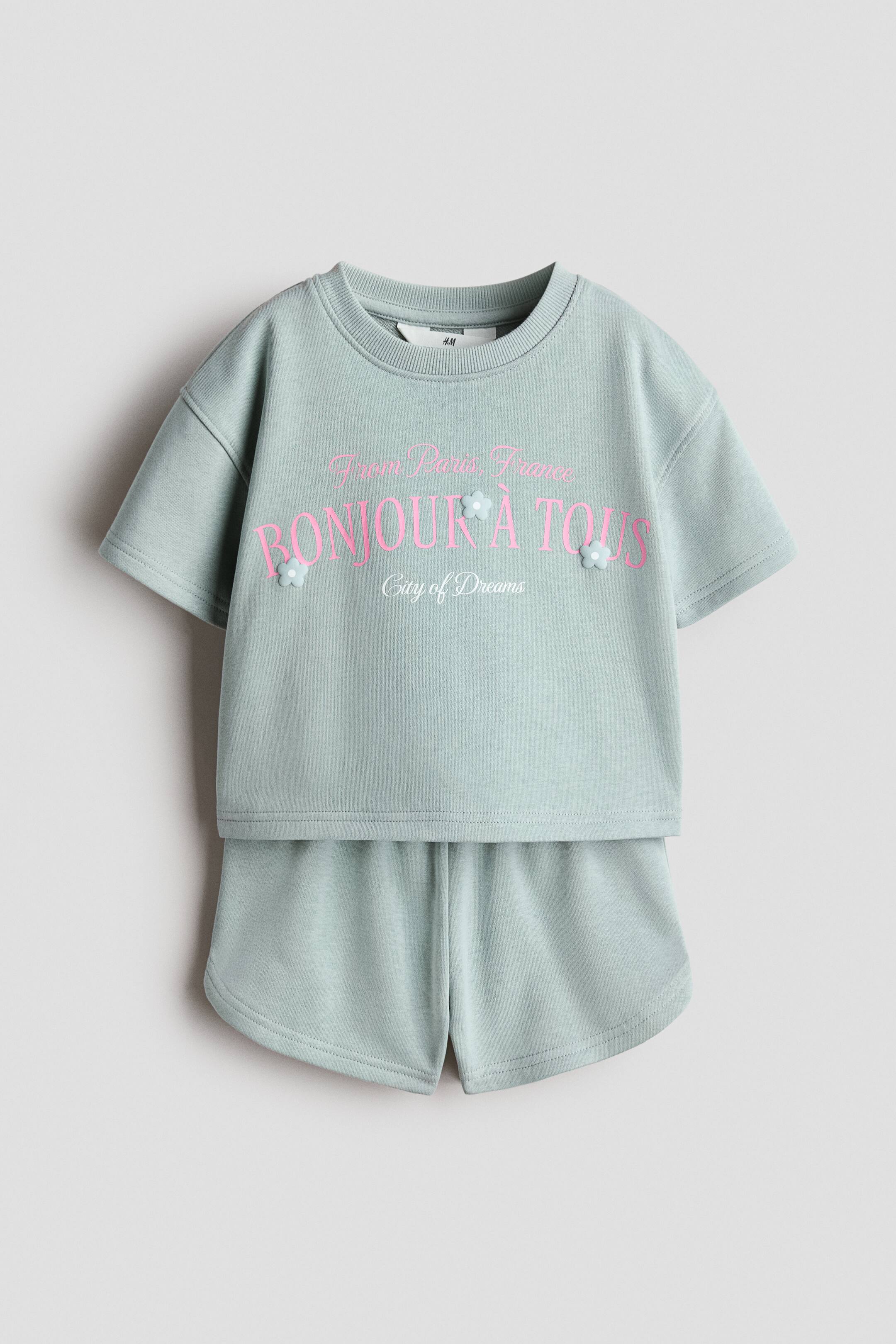 View larger image: A matching two-piece set is shown from the front, consisting of a light green short-sleeved top and coordinating shorts. The top features pink printed text reading "BONJOUR � TOUS" with small three-dimensional floral appliqués, while the shorts have a curved hem.