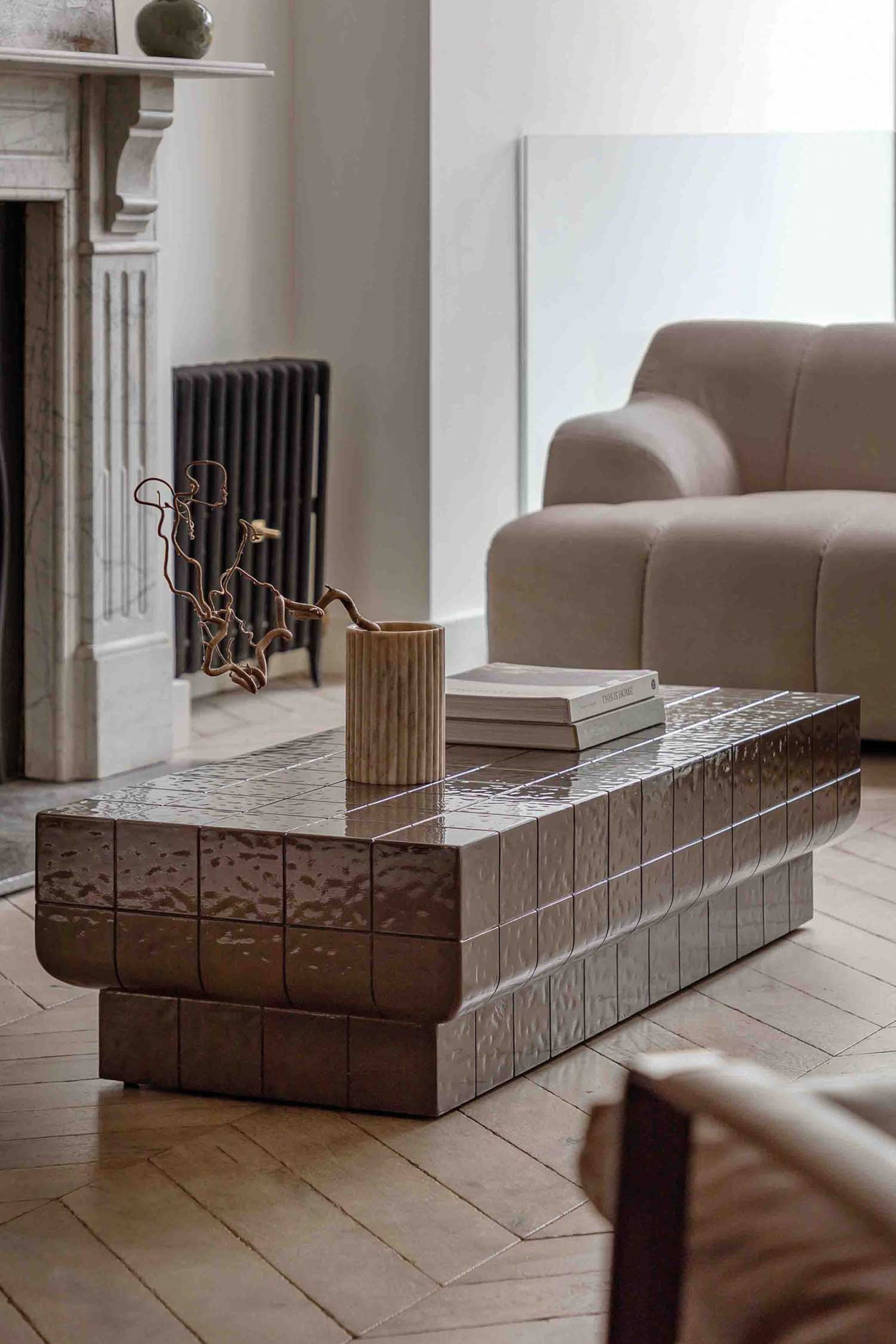 View larger image: A rectangular coffee table is depicted facing slightly towards the viewer, its top and sides adorned with glossy, dark taupe rectangular tiles. It features gently rounded corners and a recessed plinth base, creating a tiered effect. A fluted cylindrical vase and a stack of books rest on its top surface.