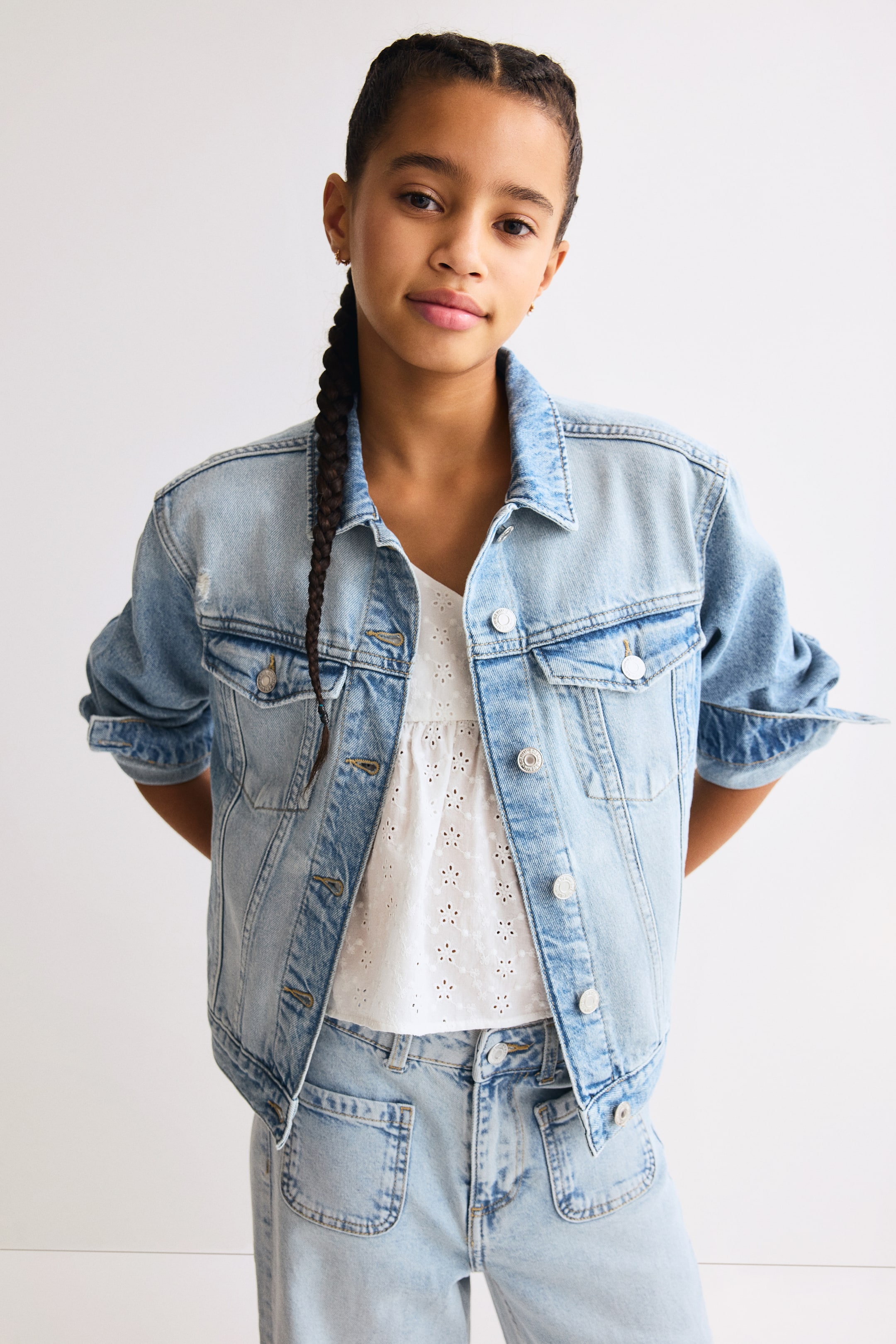 Ver imagen más grande: La chaqueta de mezclilla azul claro presenta detalles desgastados en los hombros y está diseñada con un cuello en punta y un frente abotonado. Muestra dos bolsillos con solapa en el pecho con cierres de botón y dos bolsillos inferiores tipo ojal, con sus mangas largas enrolladas en los puños. La chaqueta se lleva abierta sobre un top blanco de ojales.