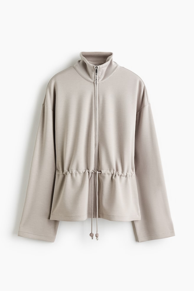 A light beige full-zip jacket showcasing a stand-up collar, dropped shoulders, and wide sleeves. It features a drawstring waist with cord stoppers, cinching the fabric. - 1