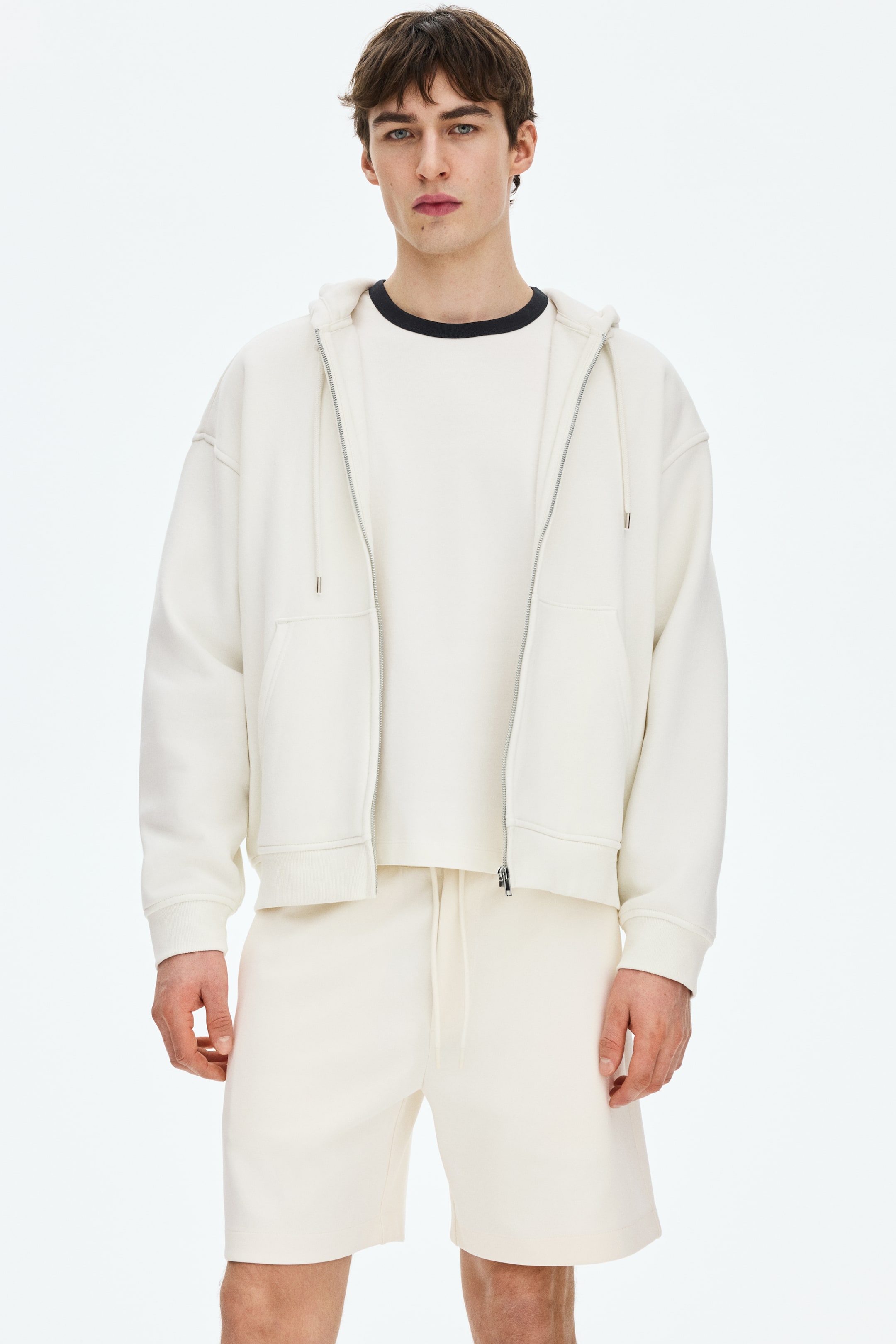 View larger image: A cream-coloured full-zip hoodie is showcased, featuring a drawstring hood, silver-toned zip, patch pockets, and ribbed cuffs and hem. It is layered over a white T-shirt with a black collar and is paired with matching cream drawstring shorts with a drawstring waist.