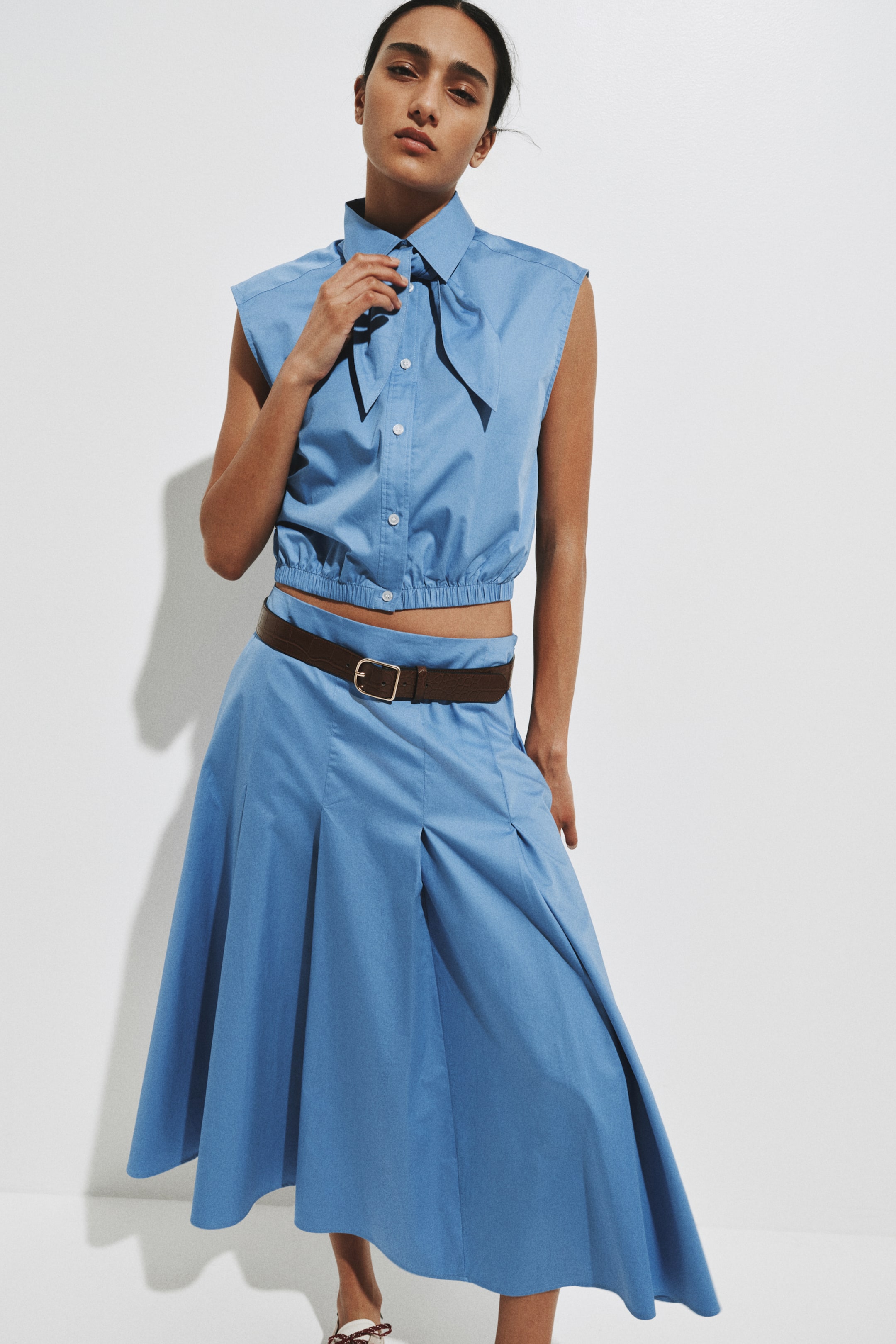 View larger image: A light blue sleeveless two-piece set features a cropped top with a pointed collar, a button-front placket, a bow tie detail at the neck, and an elasticized hem. The matching pleated midi skirt falls from the waist and is cinched by a brown belt with a gold-tone buckle.