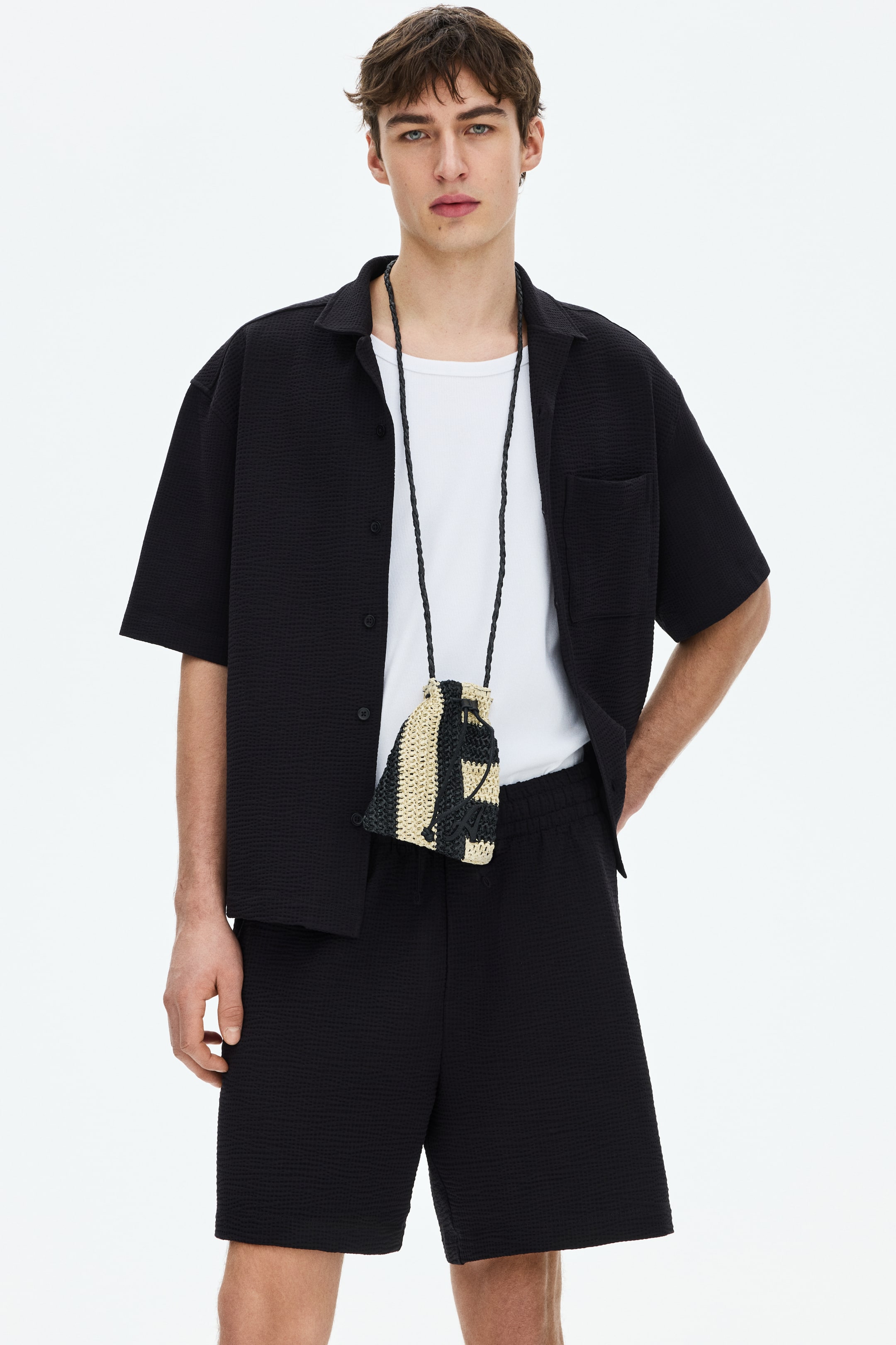 View larger image: A black textured short-sleeve shirt, featuring a camp collar and a chest patch pocket, is worn open over a white T-shirt and paired with matching black textured shorts with an elastic waistband. A small woven drawstring pouch with beige and black vertical stripes hangs from a braided strap around the neck.