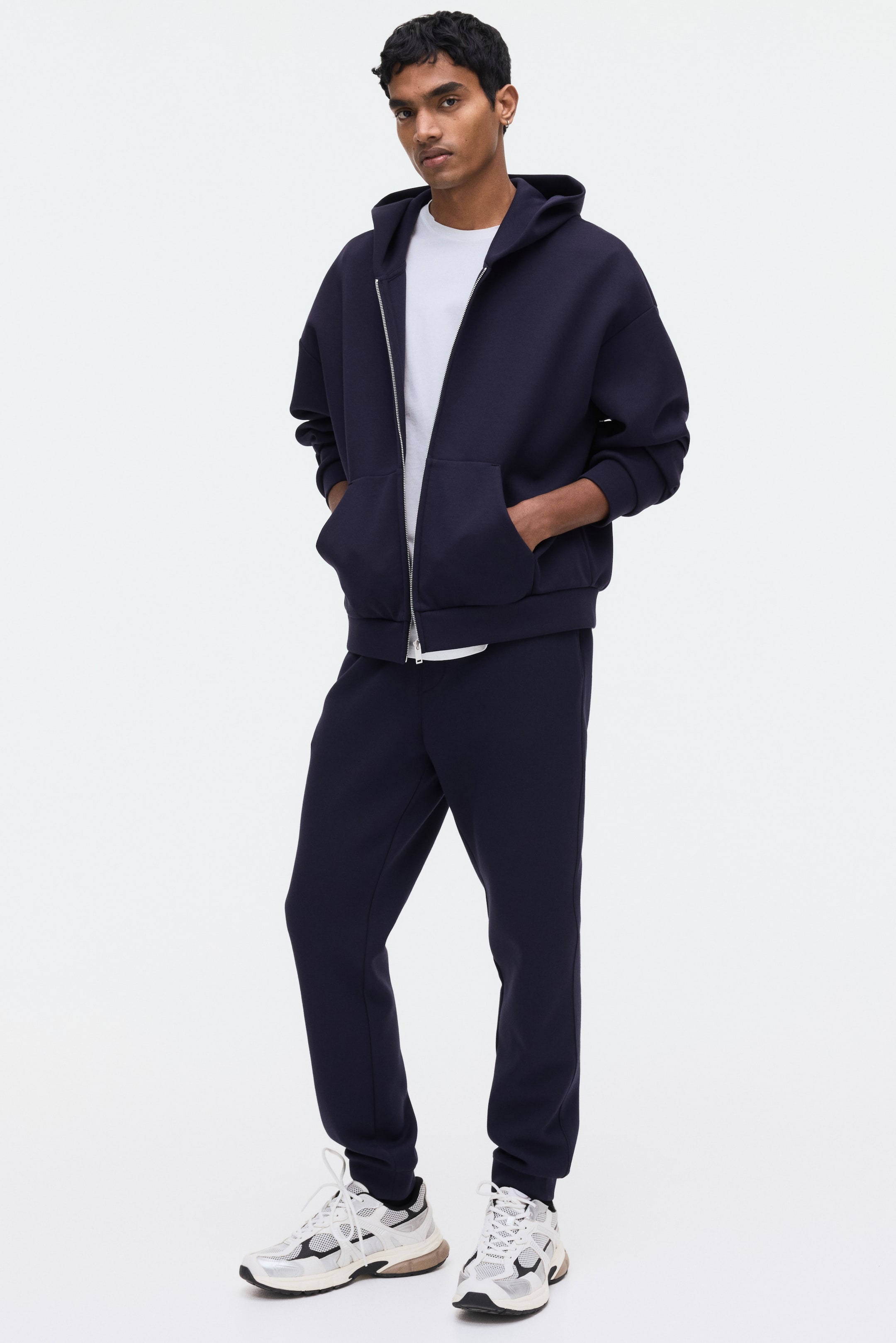 Visa större bild: Mörkblåa sweatpants med avsmalnande ben och ribbade fållar bärs av modellen, som placerar båda händerna i de diagonala sidofickorna.