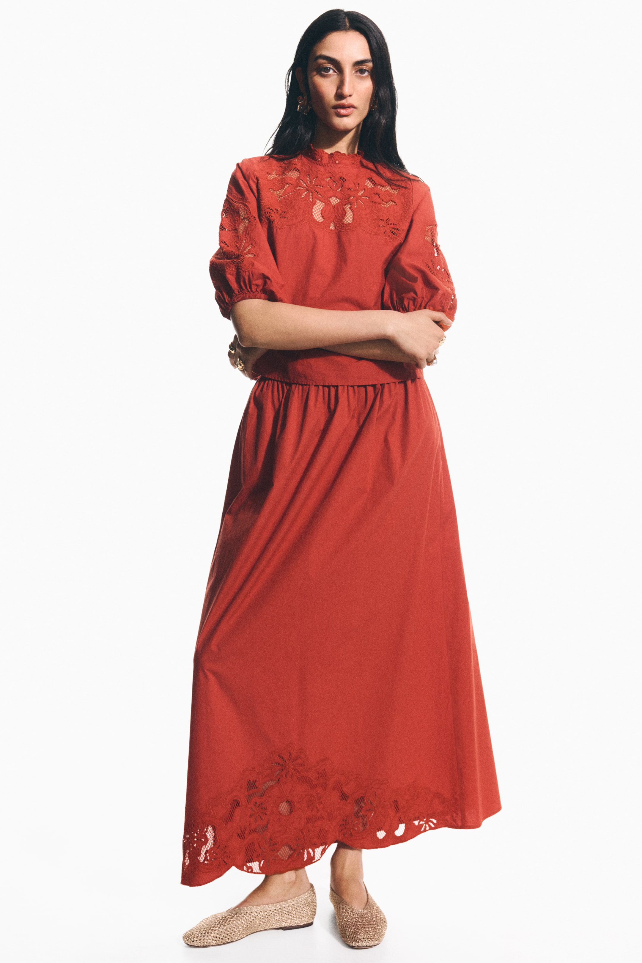 View larger image: The rust-coloured dress is showcased with a high neck, elbow-length puffed sleeves, and a flowing skirt, all highlighting intricate cutwork detailing on the yoke, sleeves, and hem.