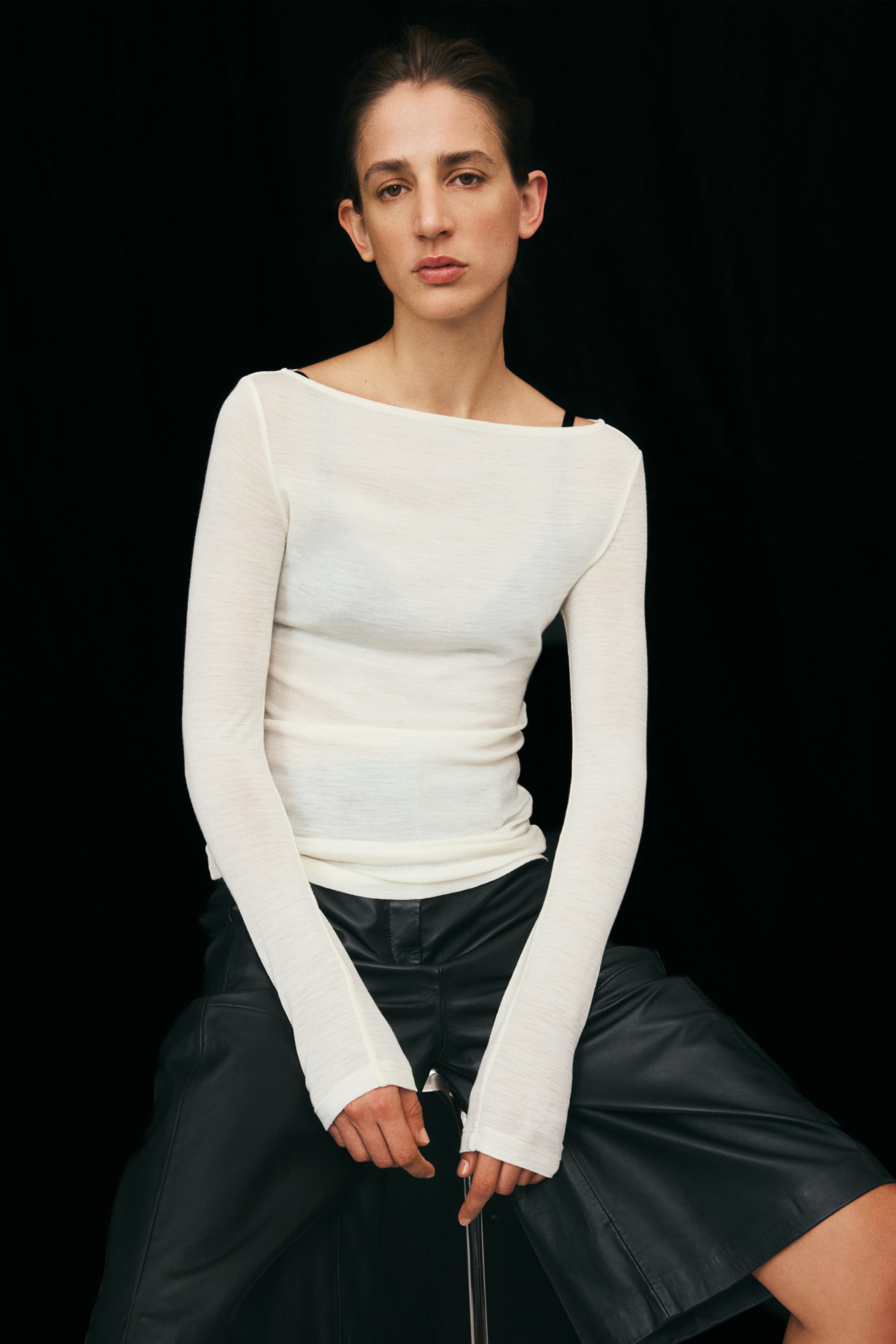 View larger image: This cream-coloured rib-knit wool top with a boat neck and long sleeves is worn by the model. The semi-sheer top is layered over a dark undergarment and paired with black leather pants.