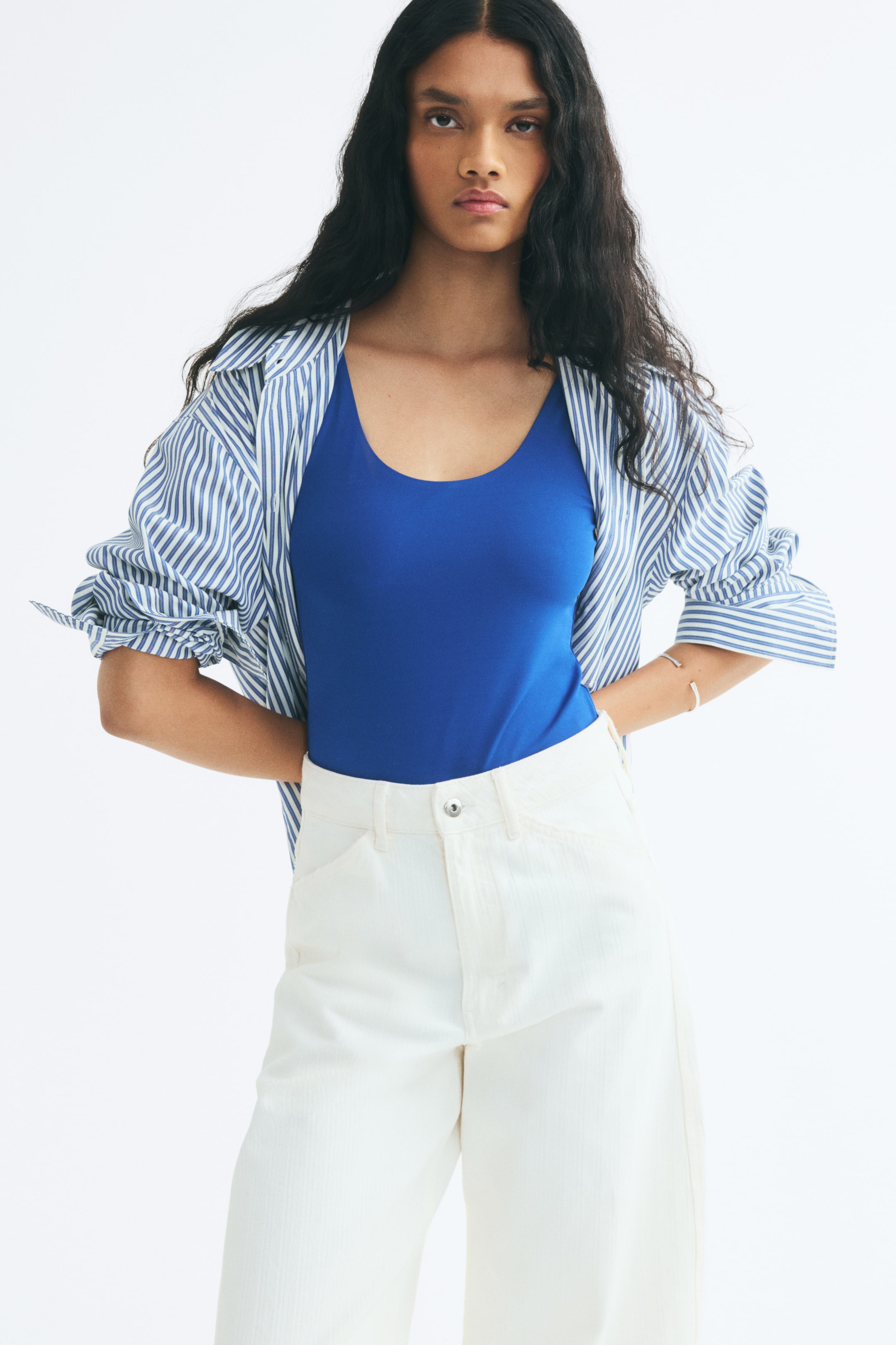Ampliar la imagen: La modelo lleva un body azul de jersey elástico con cuello redondo y tirantes anchos. El body está metido dentro de un pantalón blanco de tiro alto y se lleva debajo de una camisa abotonada abierta de rayas azules y blancas.