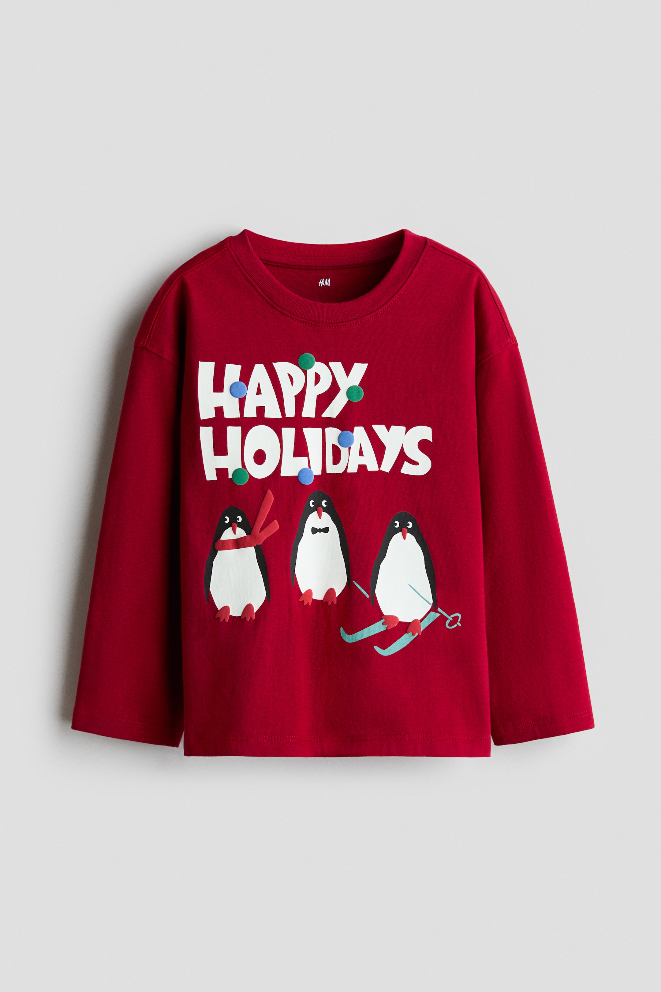 View larger image: A red long-sleeve cotton jersey T-shirt is shown from the front, featuring dropped shoulders, a round crew neckline with ribbed trim, and a printed graphic on the chest. The graphic displays the white block text "HAPPY HOLIDAYS" decorated with colourful dots above three illustrated penguins.