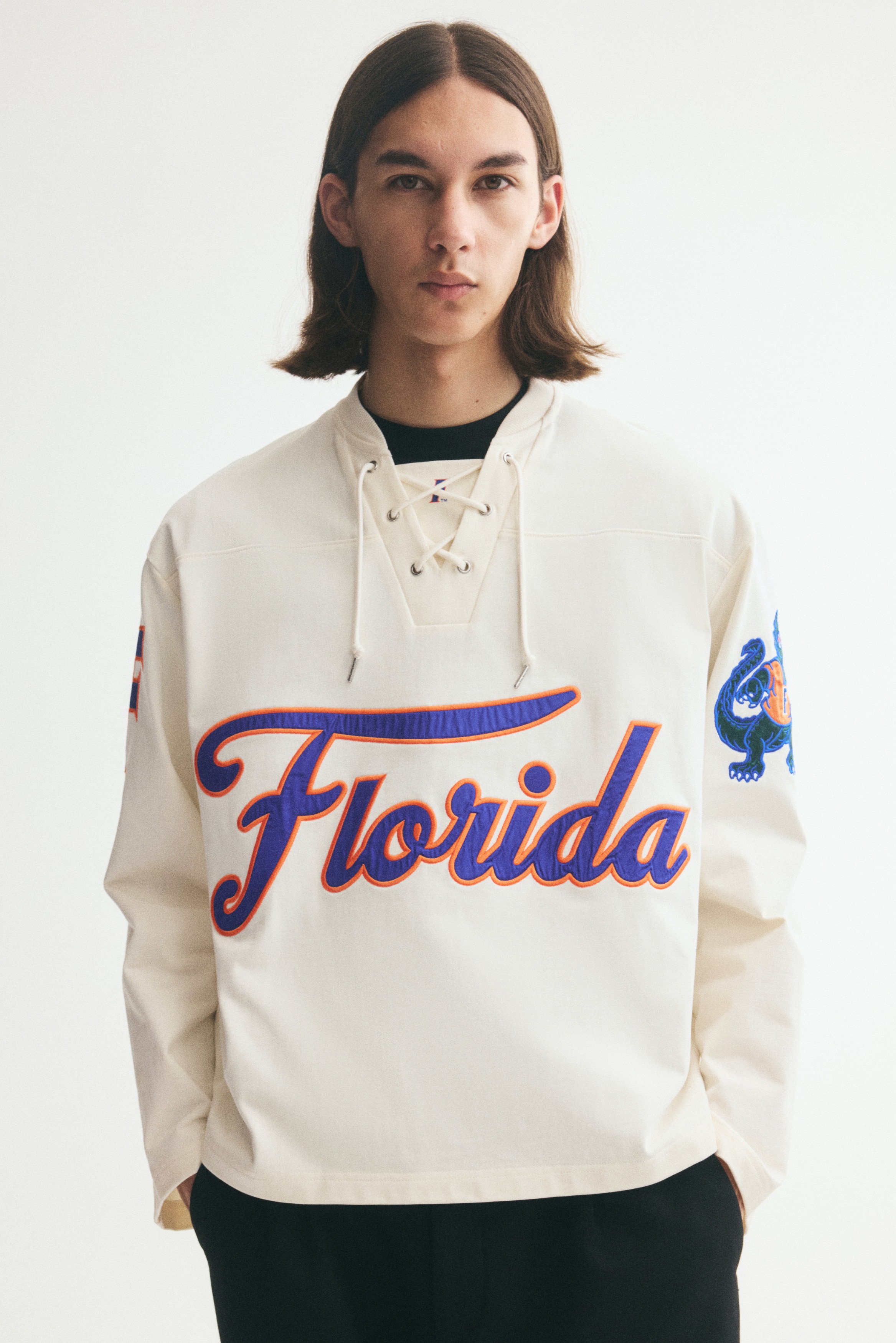 An off-white, oversized long-sleeved T-shirt is shown from a front-facing angle, featuring a lace-up V-neckline with a fabric insert and dropped shoulders. A large blue "Florida" script graphic with an orange outline is displayed across the chest, with additional collegiate motifs decorating the wide sleeves.