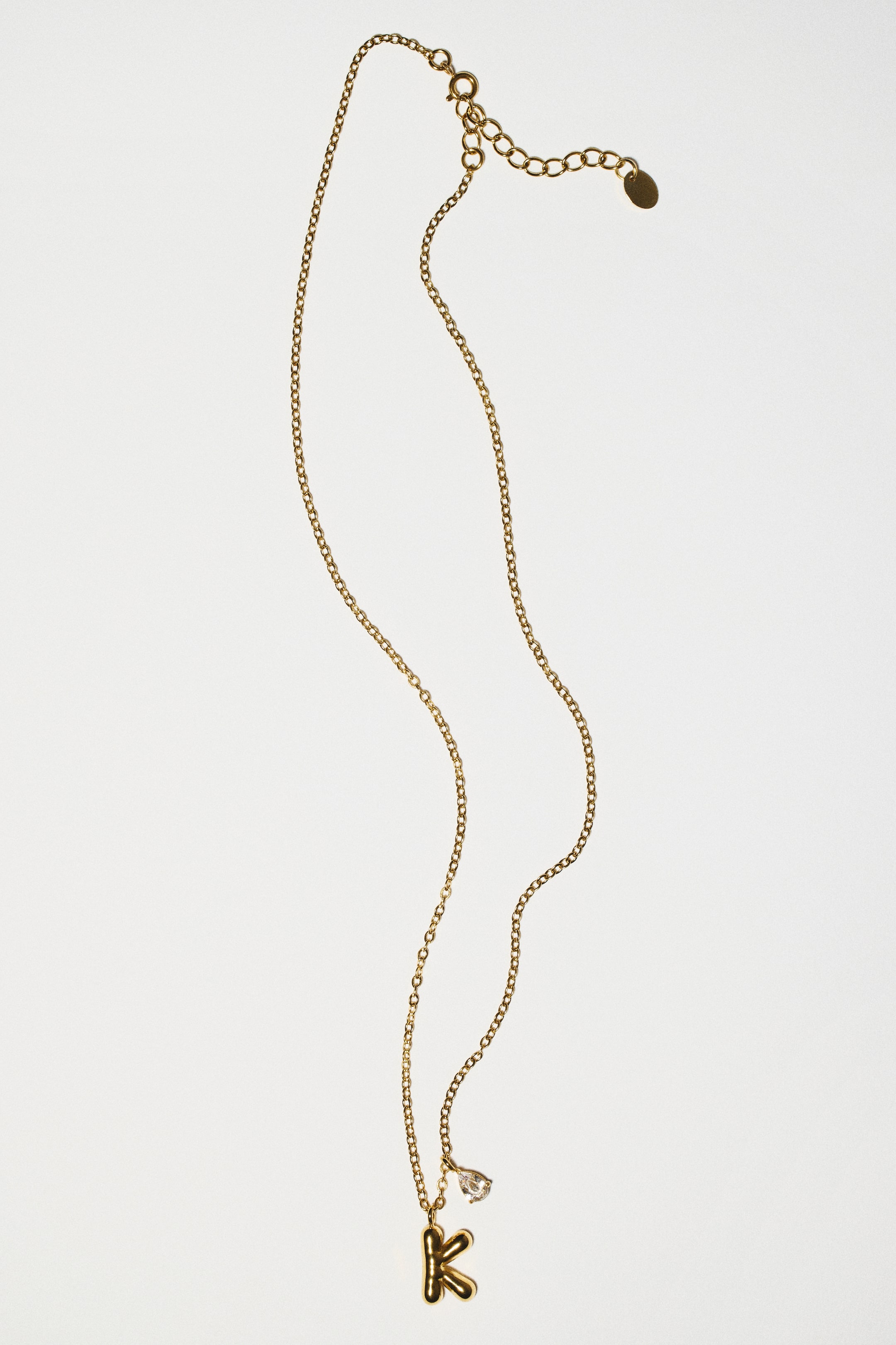 View larger image: A gold-toned stainless steel chain necklace is shown from the front, featuring a puffy letter "K" pendant alongside a small, drop-shaped clear glass rhinestone. The fine-link chain extends upwards to an adjustable section with a spring clasp.