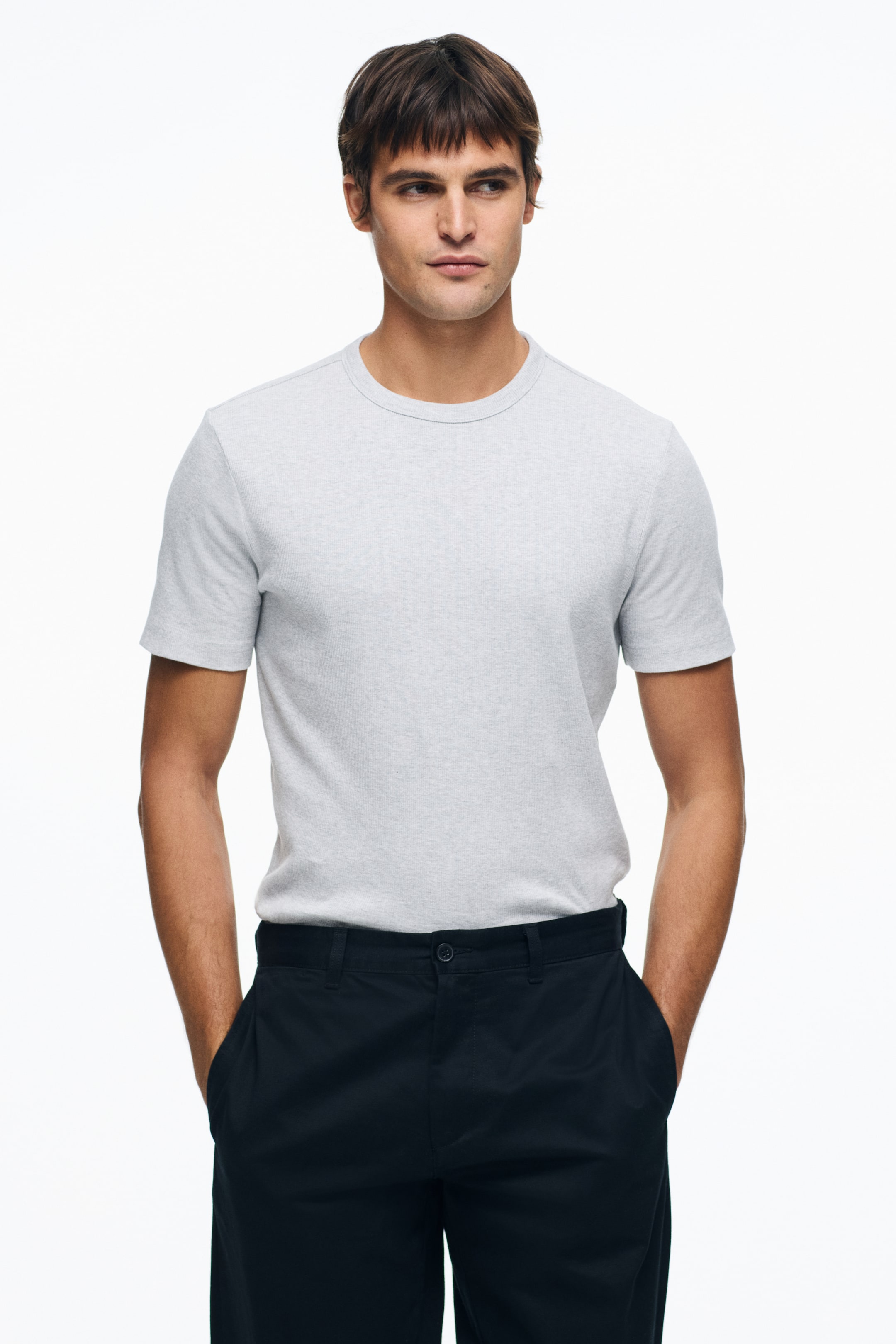 Grotere afbeelding bekijken: Een lichtgrijze marled T-shirt met ronde hals wordt gedragen, met een geribbelde kraag en korte mouwen, en is ingestopt in een donkere broek, wat het aansluitende ontwerp benadrukt.