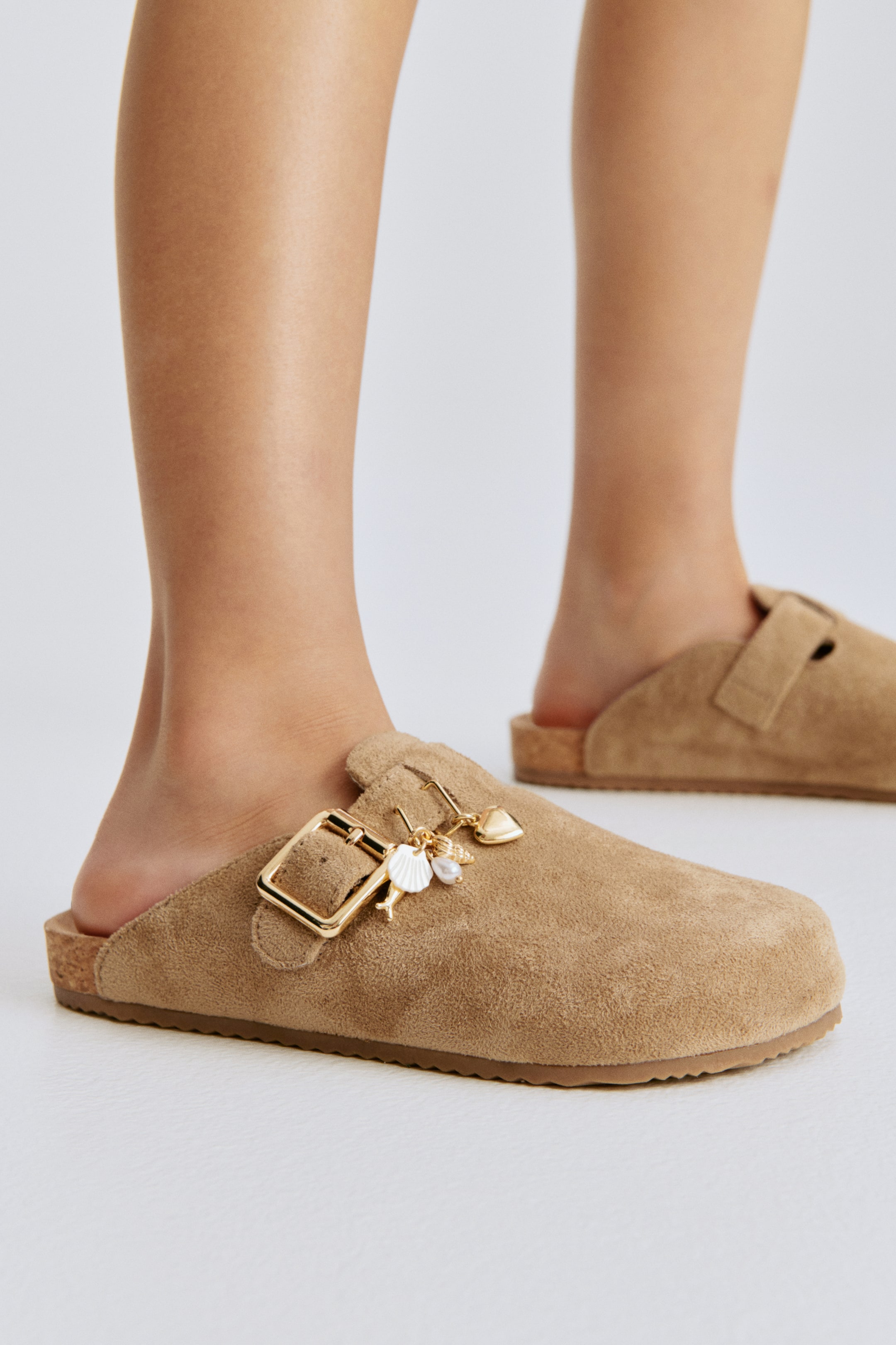Agrandir l'image: Une mule gauche en cuir fauve, fabriquée en tissu doux et duveteux, présente un bout arrondi et une bride de pied réglable avec une boucle dorée. Plusieurs breloques métalliques, dont un cœur, un coquillage et une perle semblable à une perle, pendent de la boucle, et une semelle intermédiaire façon liège est visible au-dessus d'une semelle extérieure marron.