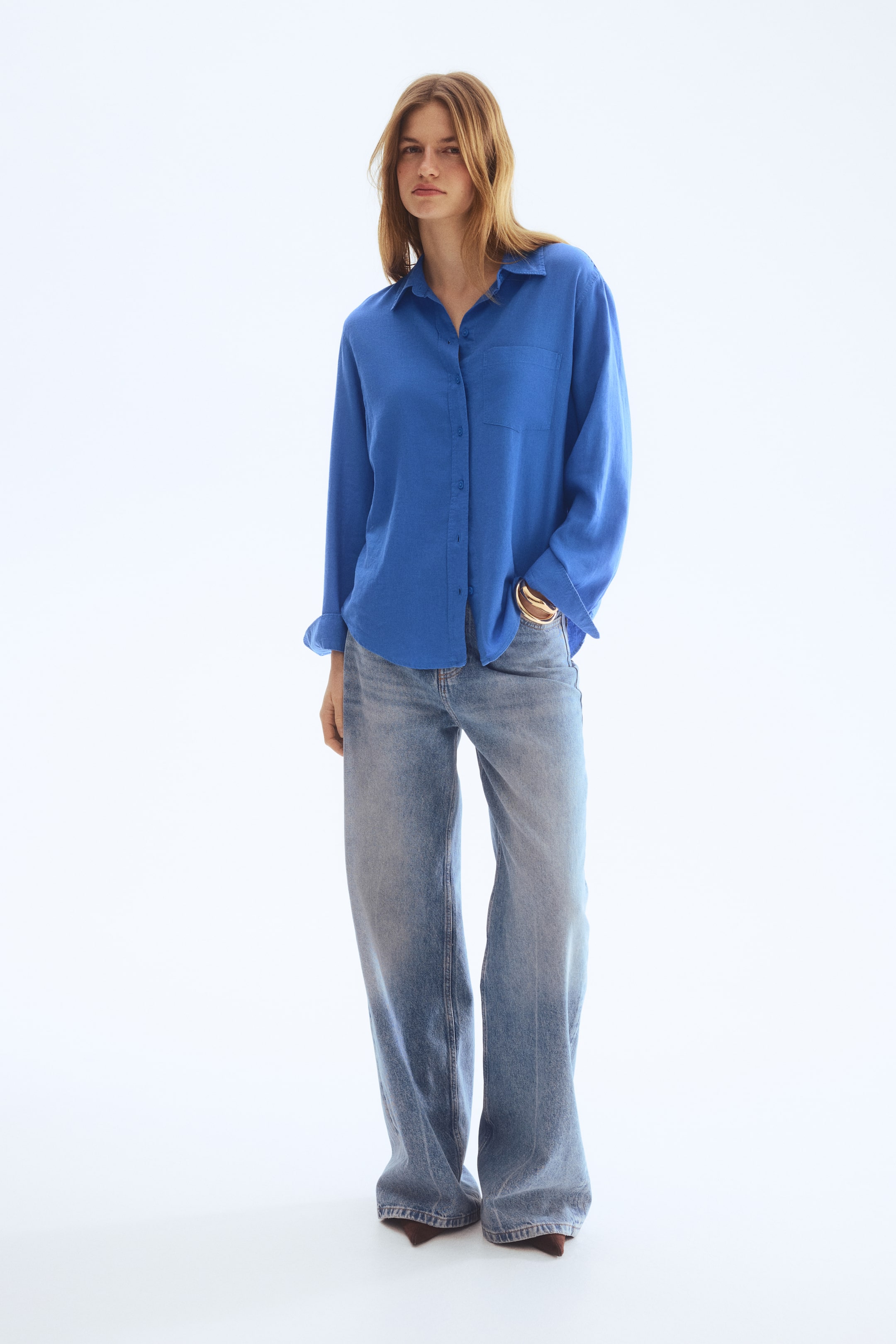 View larger image: The blue button-down shirt is facing forward, featuring a pointed collar and a left chest pocket. One long sleeve cuff is being rolled up, revealing a gold-coloured bracelet, and the shirt is complementing light wash, wide-leg jeans that are showcasing a relaxed fit.