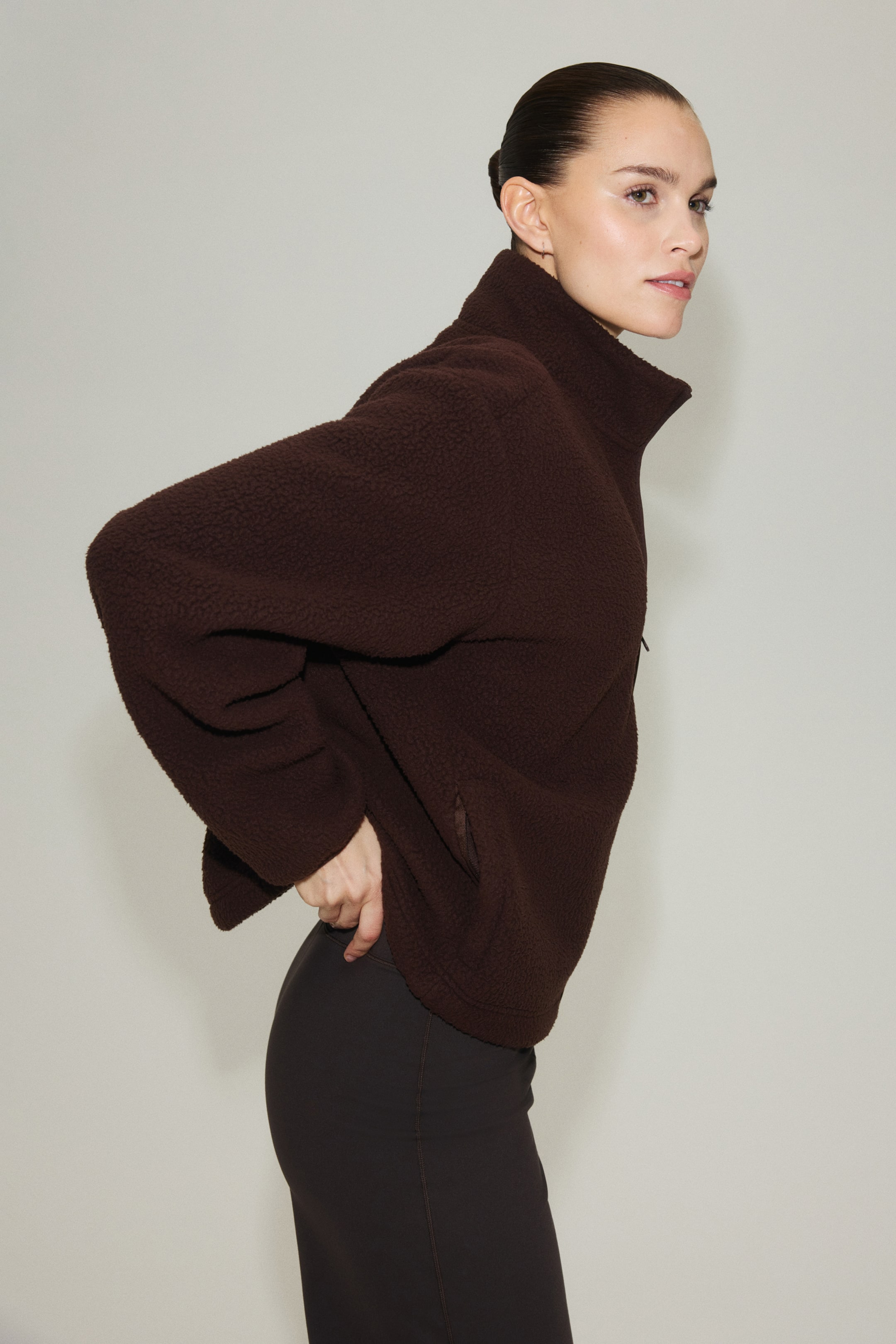 Visa större bild: En brun fleece-tröja visas från en snett bakifrån vinkel, med en hög krage med en delvis dragkedja, långa ärmar och sidofickor.