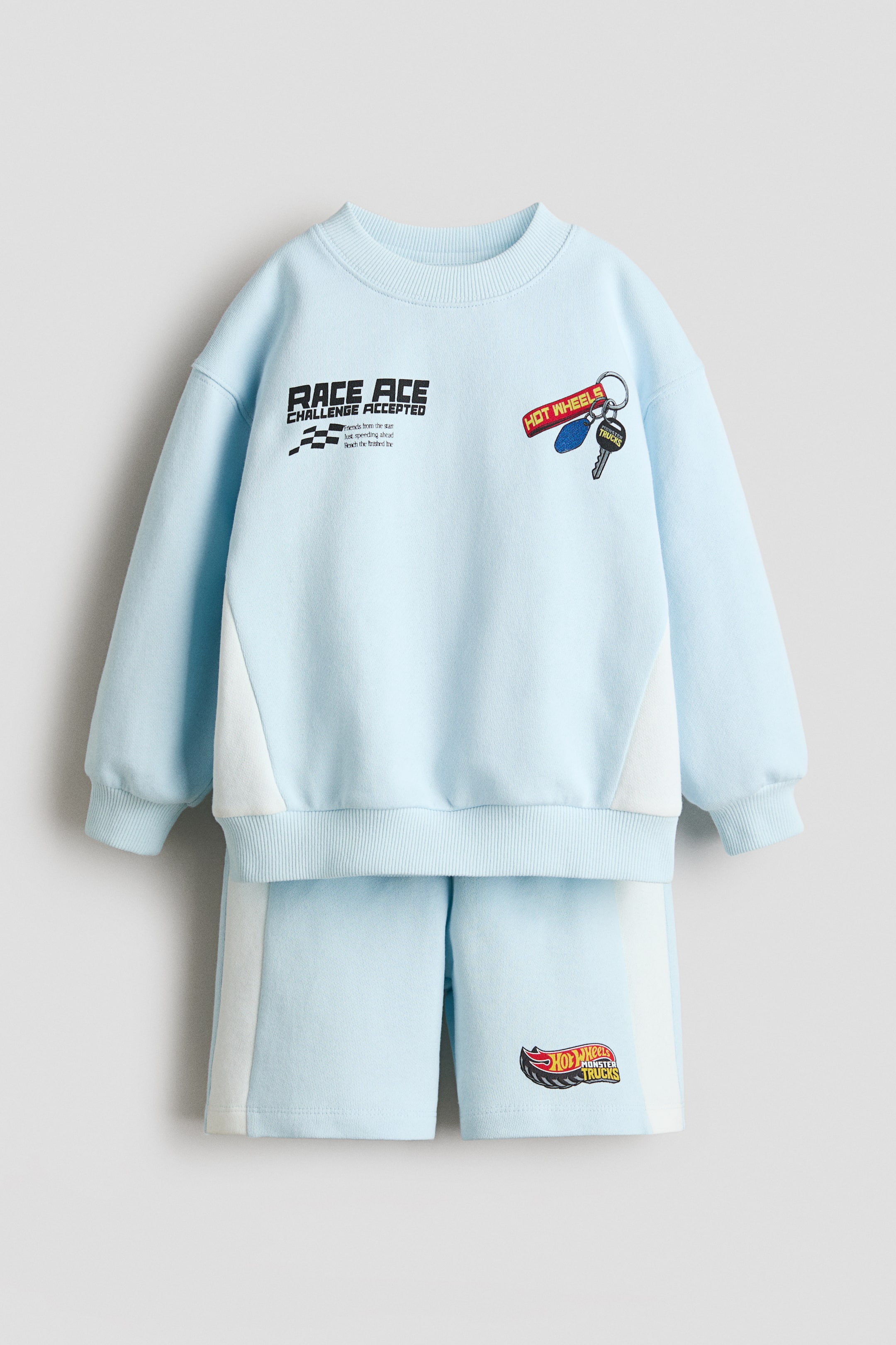 View larger image: A light blue cotton sweatshirt set featuring a long-sleeved top with dropped shoulders and matching shorts, both displaying white side panel accents. The top showcases "RACE ACE" text and a "HOT WHEELS" keychain graphic on the front, with ribbed detailing on the neckline, cuffs, and hem. The shorts present a "HOT WHEELS MONSTER TRUCKS" logo on the front left leg.