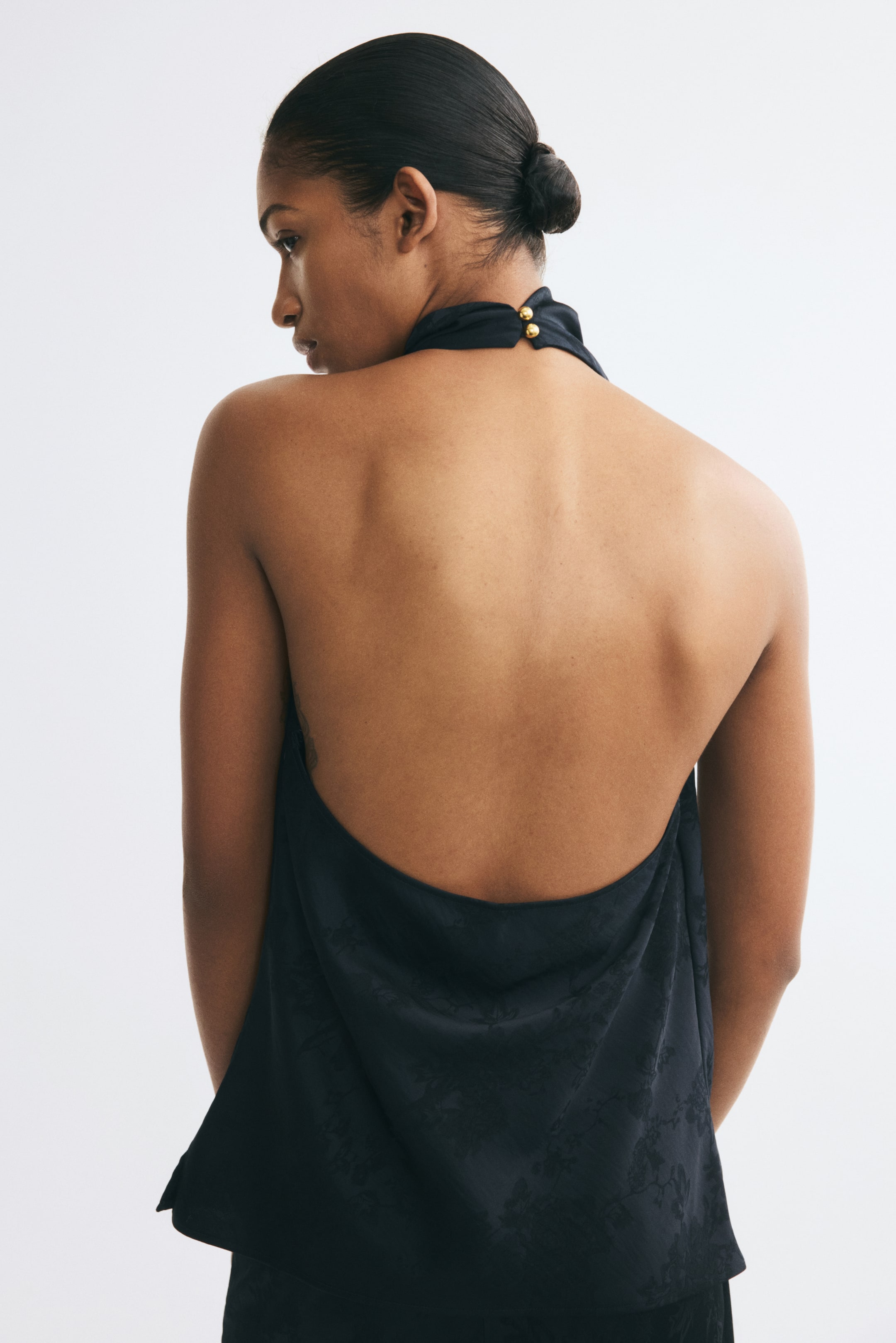 View larger image: The model is wearing a black halterneck top in a floral jacquard weave, highlighting its low open back. The top features a wide halter strap fastened behind the neck with two gold-toned buttons.