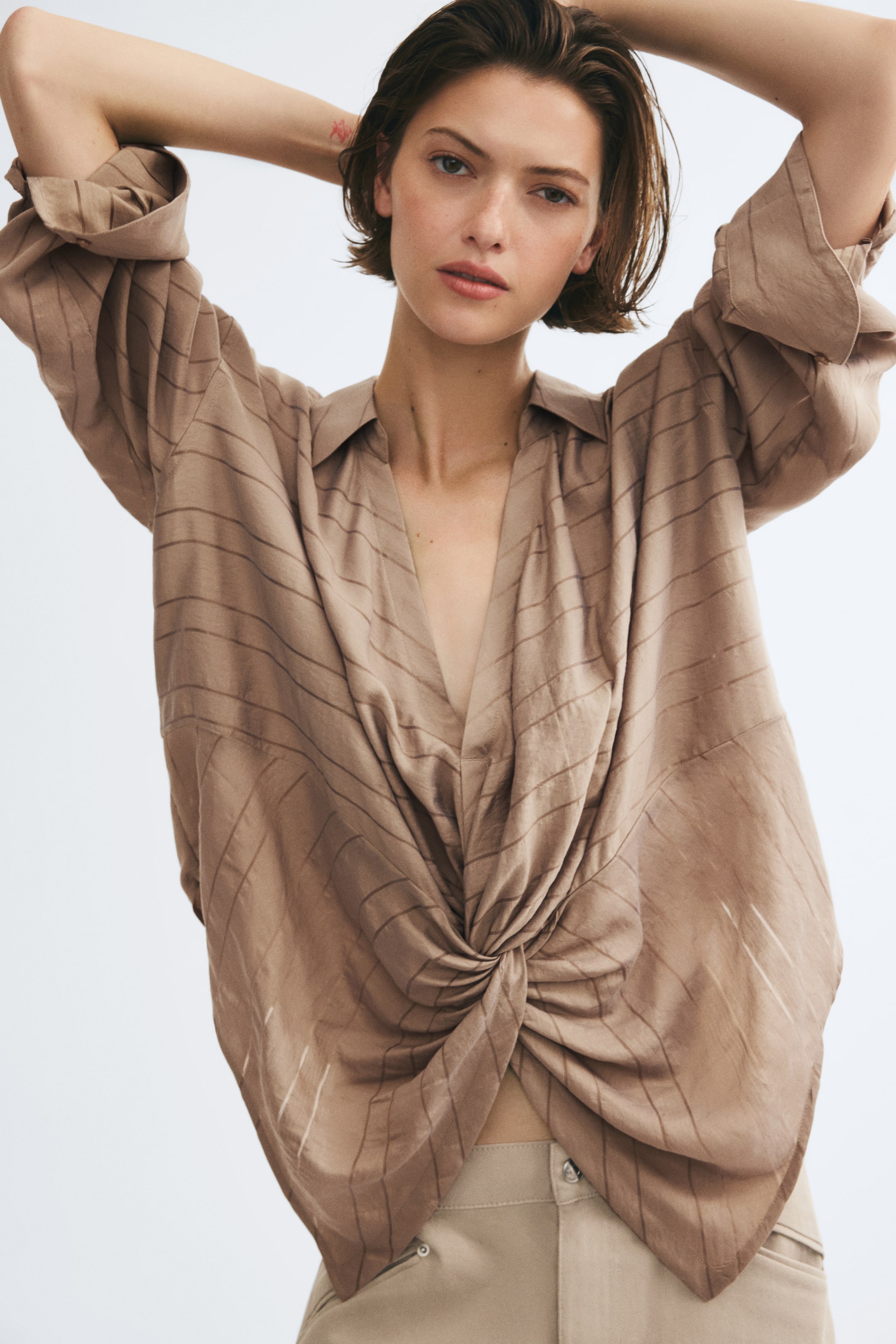 Ver imagen más grande: La modelo lleva una camisa a rayas de color marrón claro con cuello abierto y detalle frontal retorcido. La camisa, de corte holgado, se combina con pantalones beige mientras la modelo posa con ambas manos levantadas detrás de la cabeza.