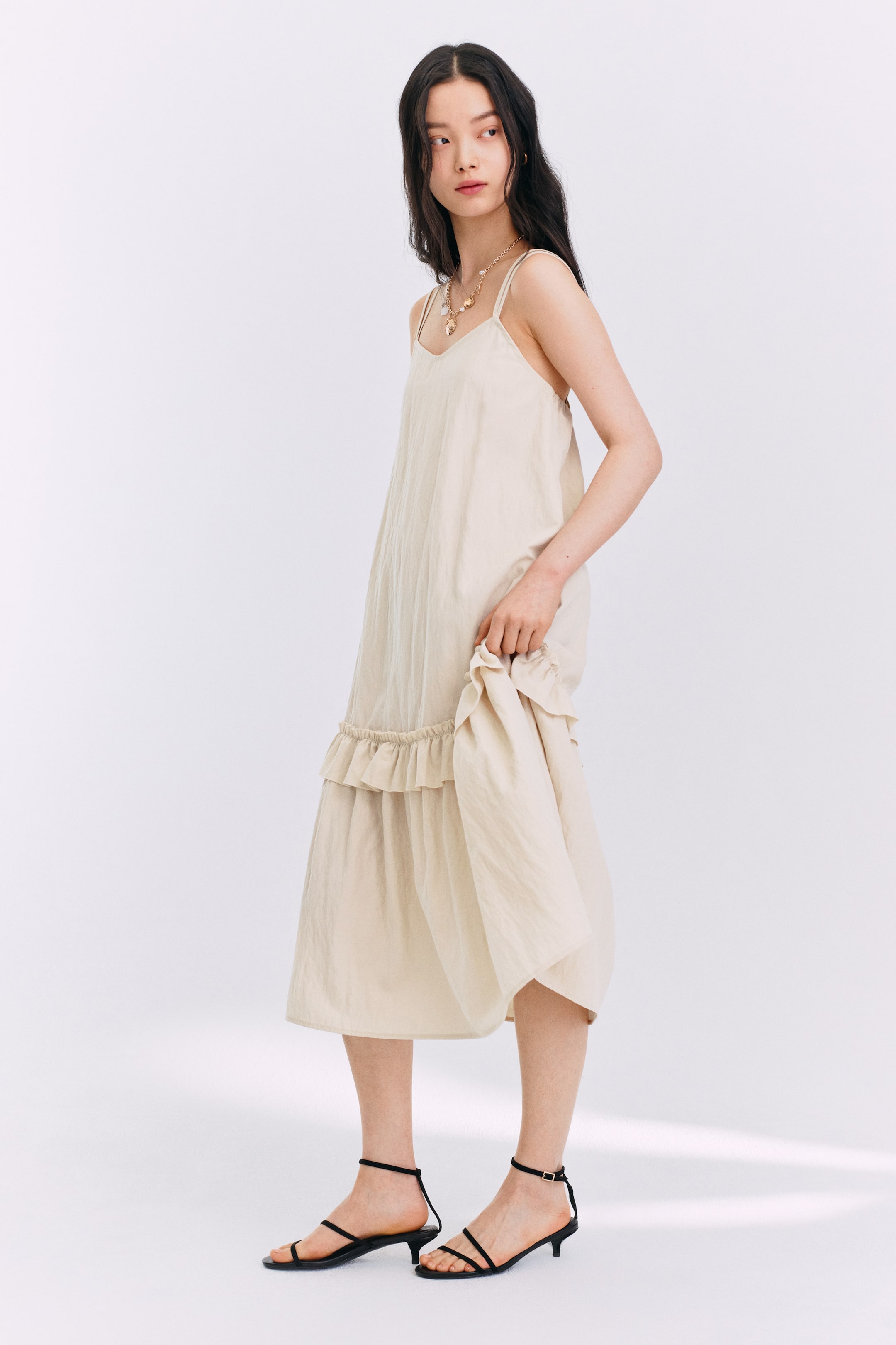 拡大画像を表示: 薄いベージュのミディドレスは、細いスパゲッティストラップとVネックラインが特徴です。このゆったりとしたドレスは、裾の上にフリルのティアがデザインされ、膝下まで落ちます。