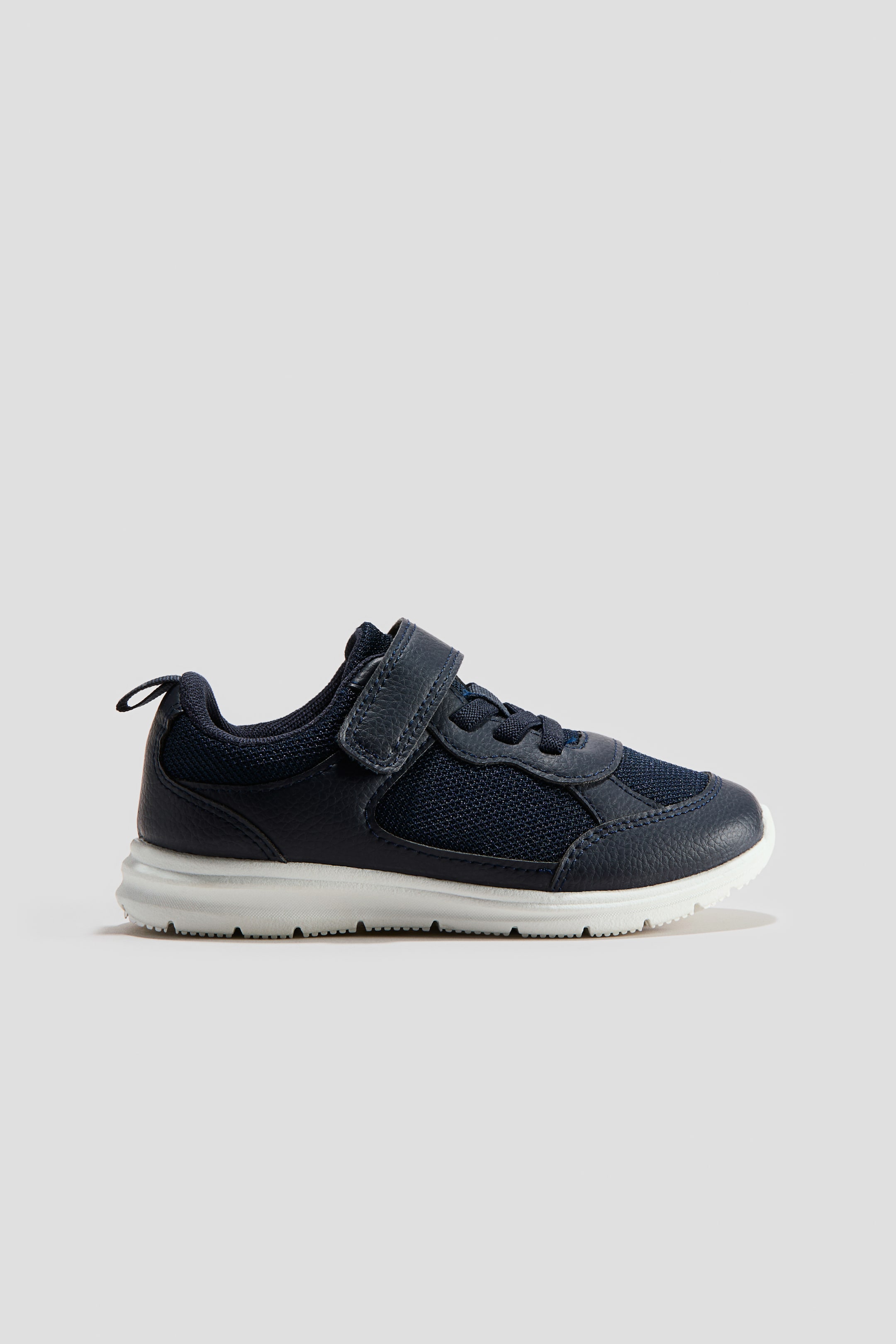 View larger image: A navy blue trainer is shown in a side view, featuring coated fabric with mesh details, a hook-and-loop tab at the top, elasticated lacing at the front, and a grosgrain loop at the back.