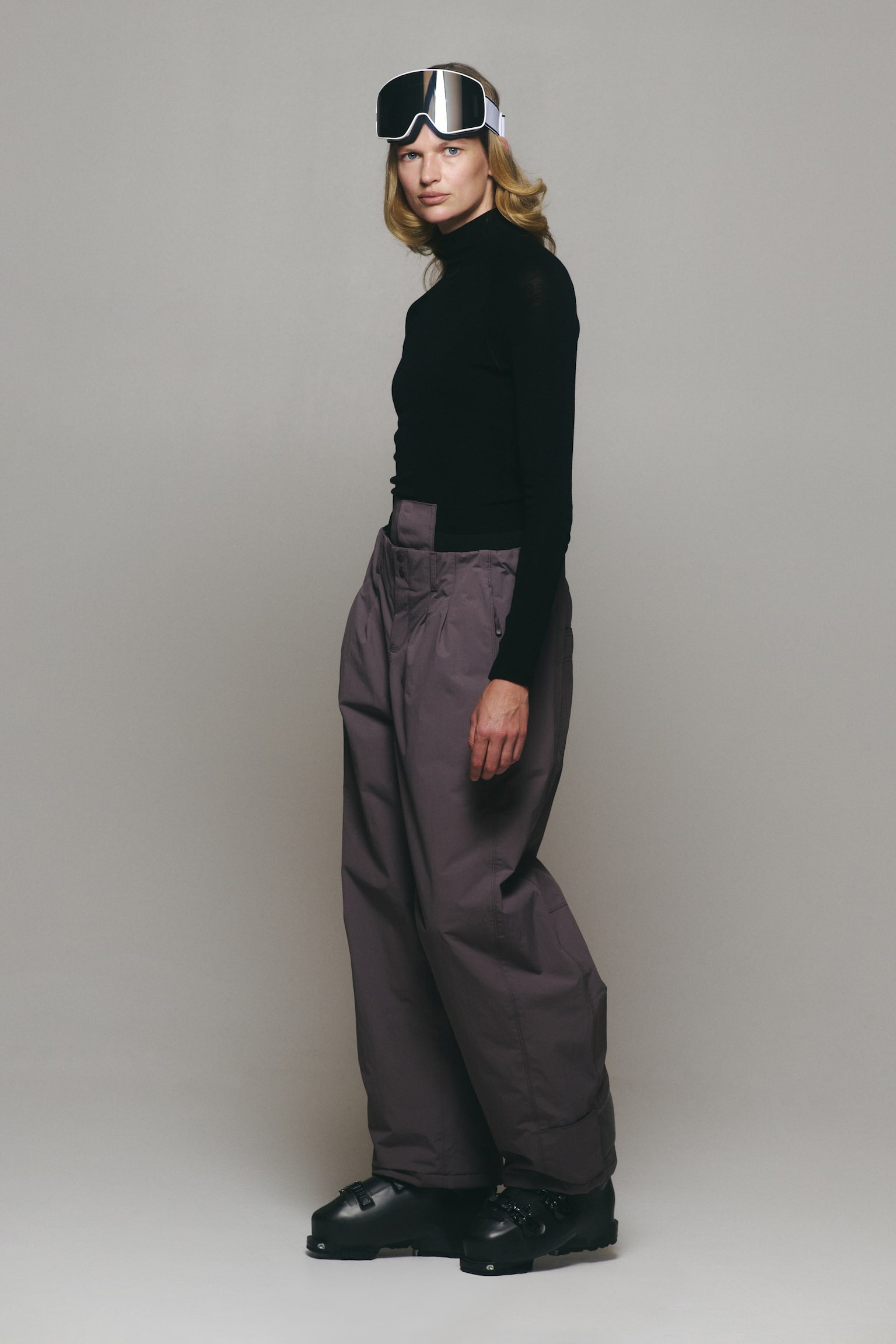 The model is wearing muted purple wide-leg ski trousers, featuring a high-waisted fit and a relaxed silhouette. The trousers are styled with a black long-sleeve turtleneck, white-framed ski goggles, and black ski boots. - 1