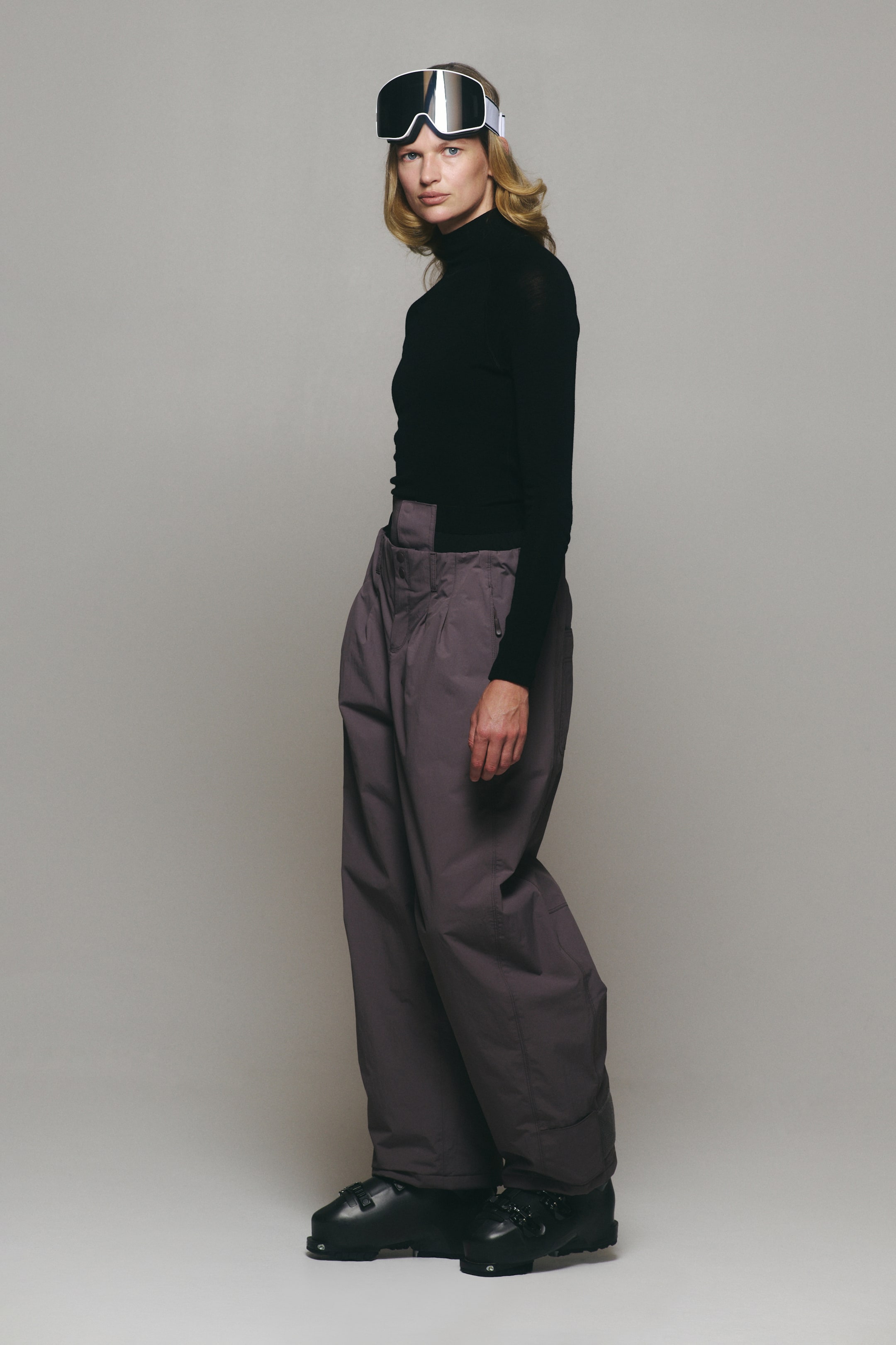 View larger image: The model is wearing muted purple wide-leg ski pants with a high-waisted fit and a relaxed silhouette. The pants are styled with a black long-sleeve turtleneck, white-framed ski goggles, and black ski boots.