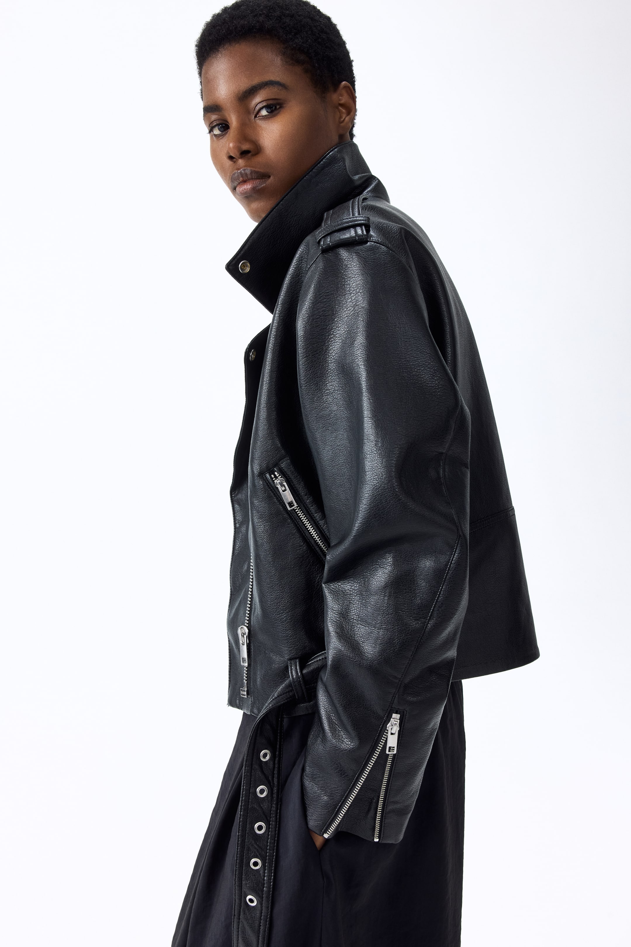 Grotere afbeelding bekijken: Een zwart leren motorjack wordt gepresenteerd vanuit een zij-voorhoek. Het heeft een gestructureerde afwerking, een asymmetrische voorrits, een opstaande kraag met een drukknoop op de linker revers en schouderepauletten. Een schuin geritste zak, lange mouwen met ritssluitingen aan de manchetten en een kleine verstelbare riem met gesp aan de zijkant zijn ook zichtbaar.